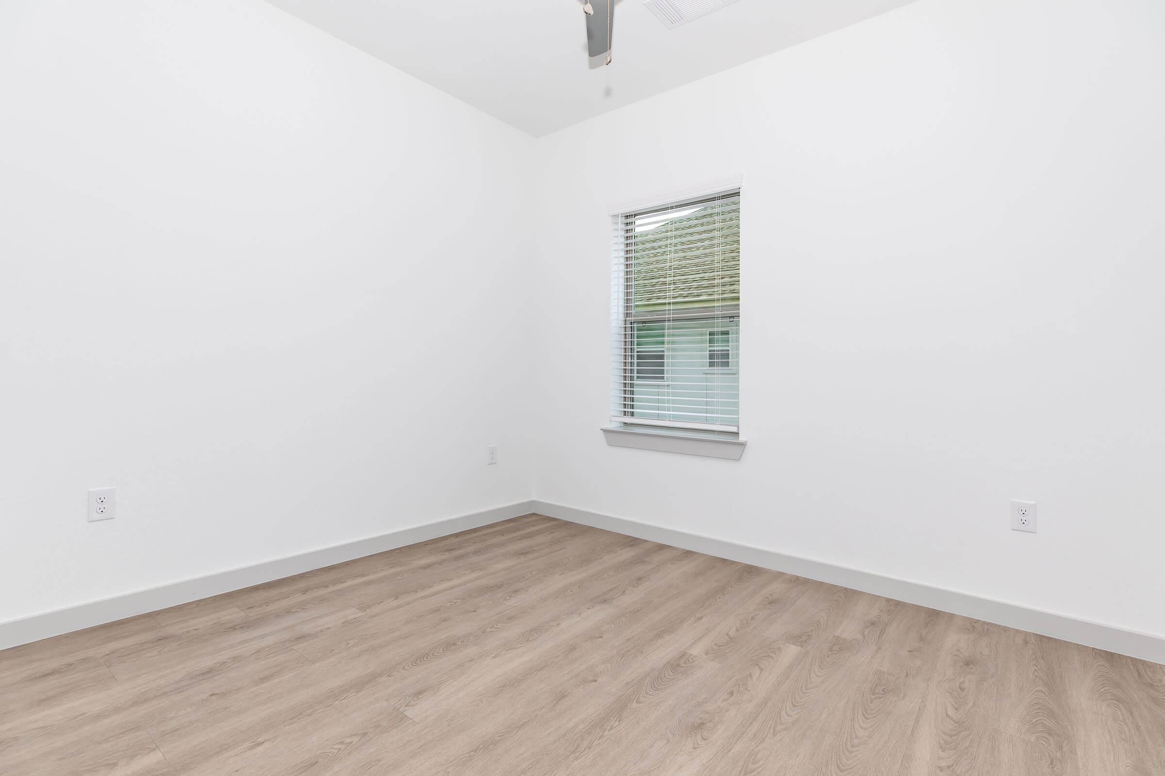 A bright, empty room with white walls and a light-colored wooden floor. There is a window with blinds on one wall, allowing natural light to enter. The ceiling features a ceiling fan, and the overall atmosphere is minimalistic and clean.