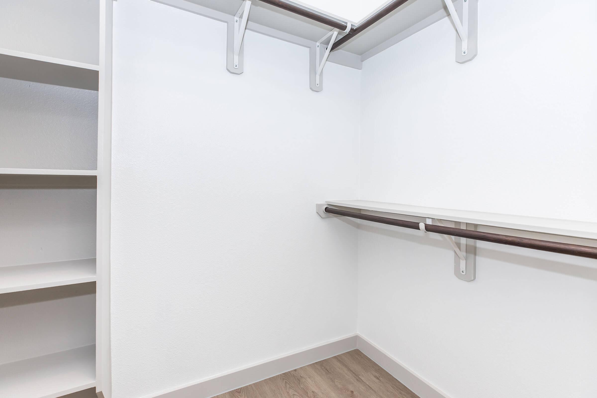 A clean, empty closet featuring white walls, a wooden floor, and open shelving. Two metal rods for hanging clothes are mounted on the wall, with a shelf above and a larger shelf on the left side. The overall design is minimalist and spacious, ideal for organizing belongings.