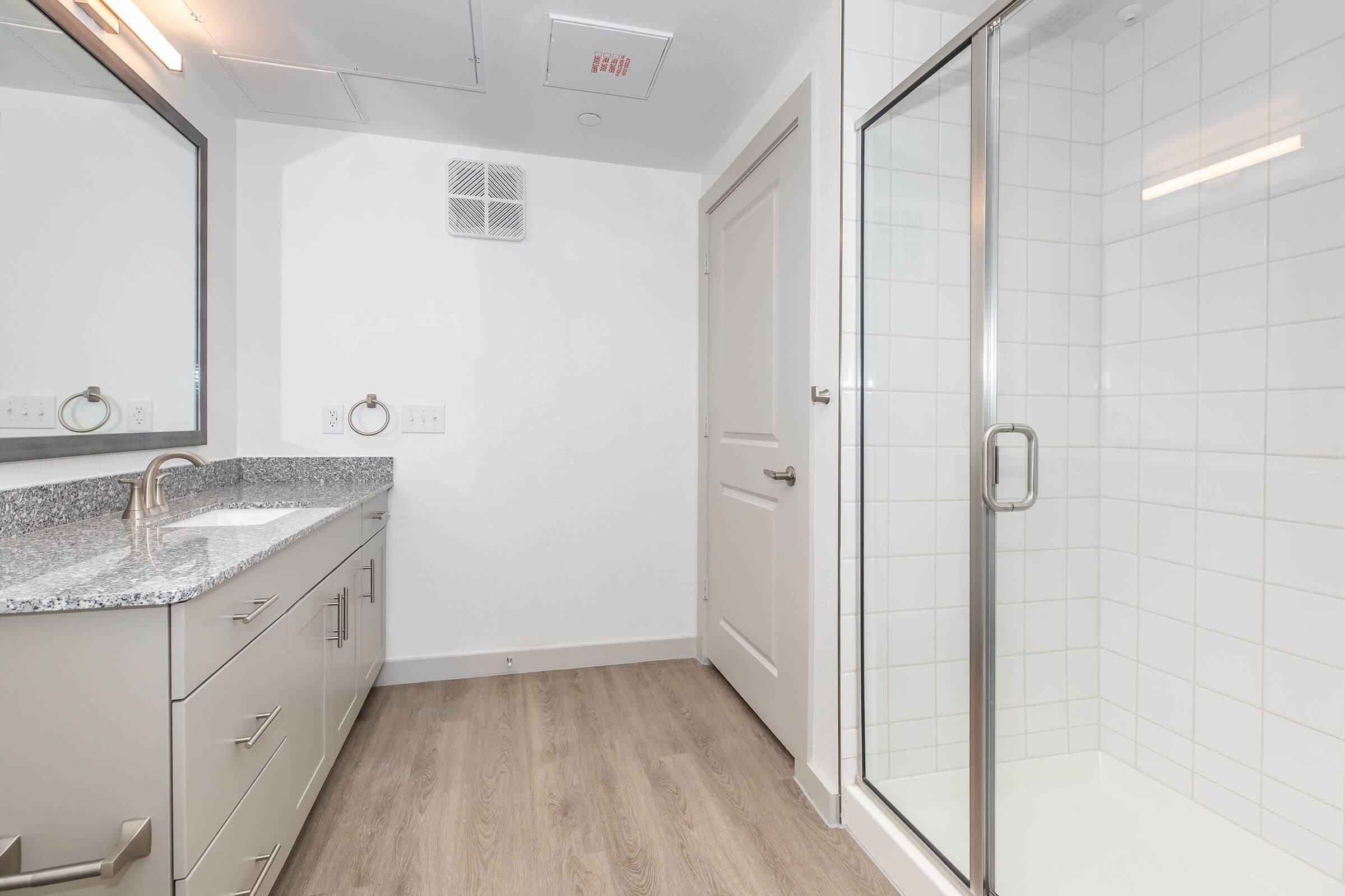 Modern bathroom featuring a walk-in shower with glass doors, a double vanity with a dark granite countertop, and two large mirrors. The floor is covered with light-colored wood-like tiles, and the walls are painted a clean white, creating a bright and spacious atmosphere.
