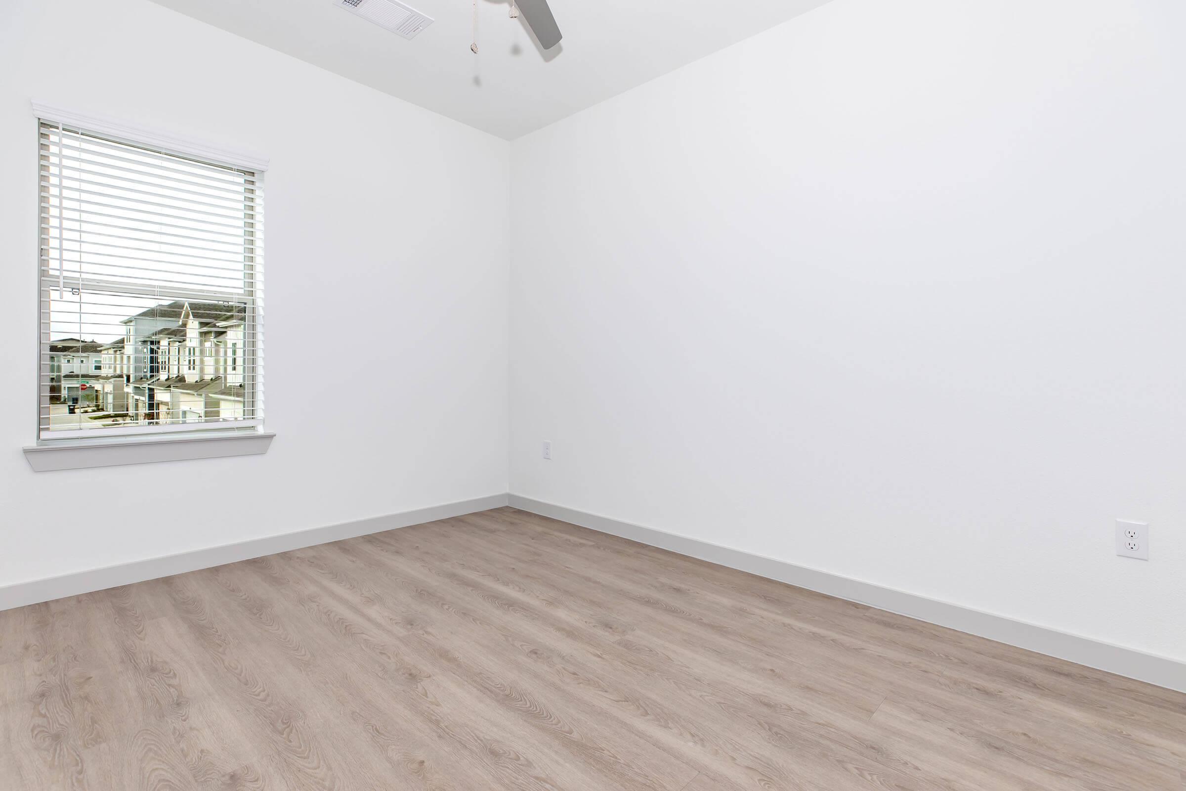 Empty room with white walls and light-colored laminate flooring. A window with blinds allows natural light in, revealing a view of the exterior. The space features a ceiling fan and is uncluttered, providing a minimalist aesthetic.