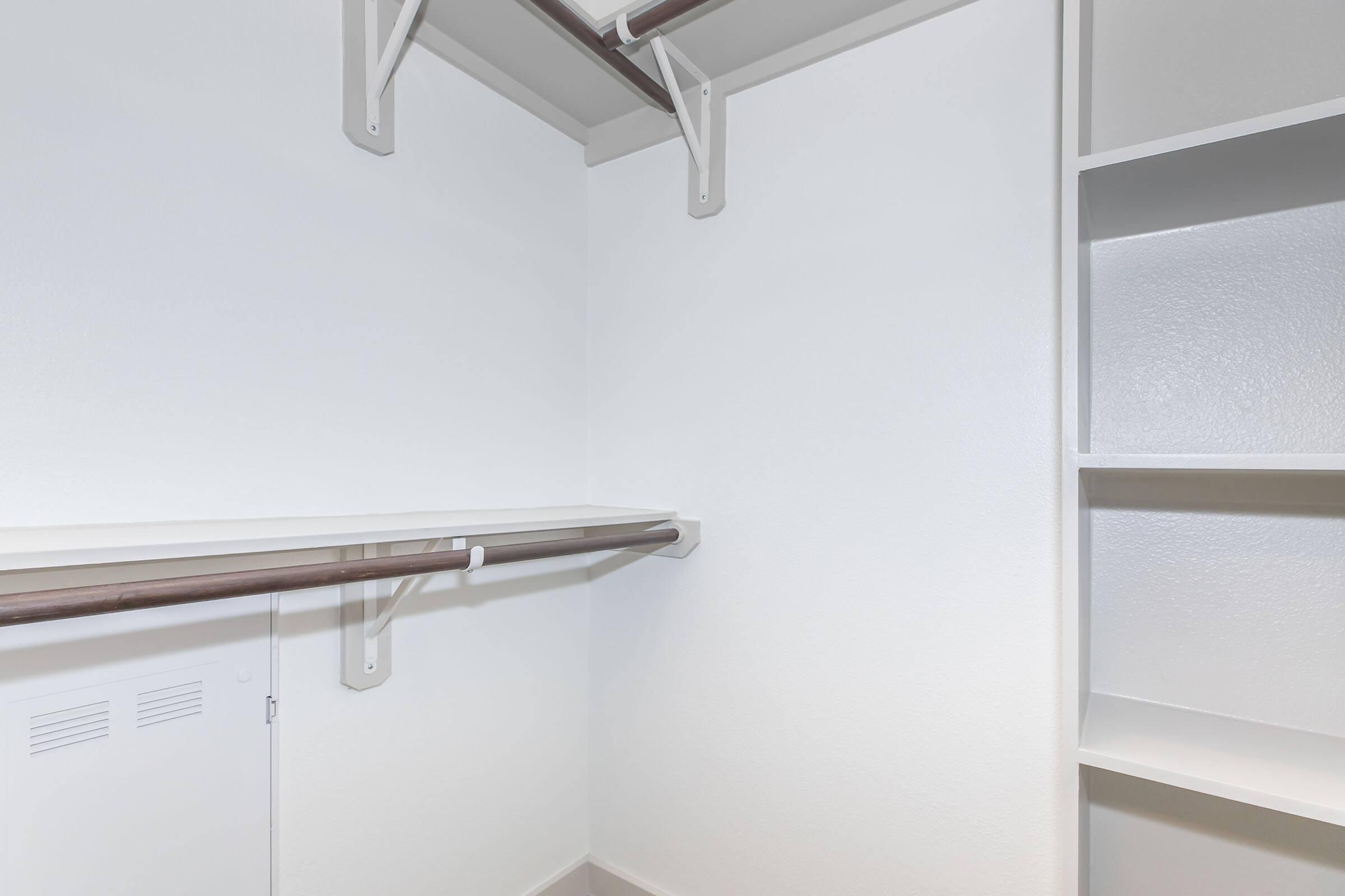 An empty closet with white walls and a light-colored floor. It features a single shelf along one side and hanging rods above, providing storage space. The opposite side has built-in shelves for additional organization. The overall design is clean and minimalistic.