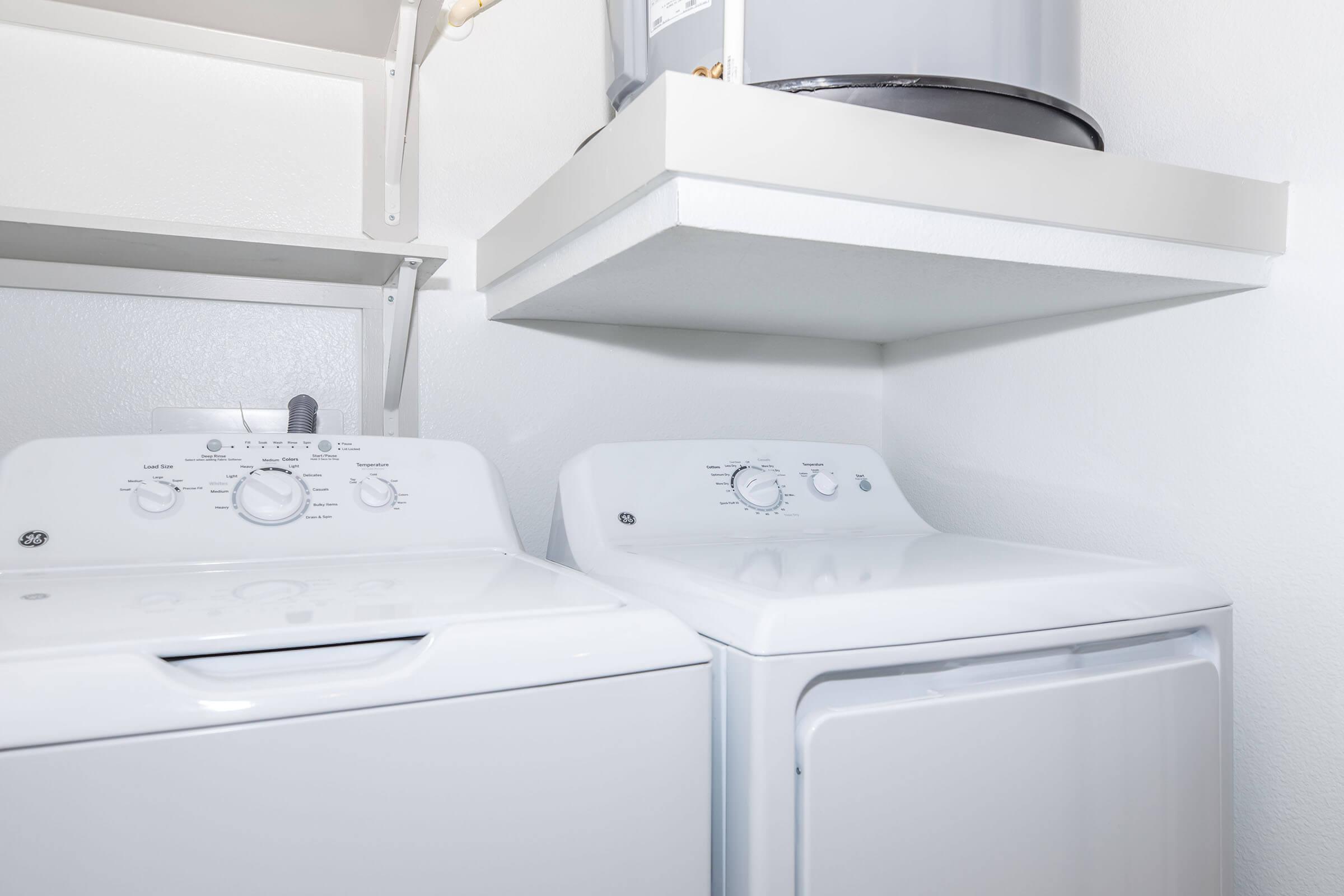 A laundry area featuring a white washer and dryer side by side. The washer has control knobs on the top, while the dryer displays a similar design. Above them is a shelf, and a gray storage container is visible. The walls are painted white, creating a clean and organized look.