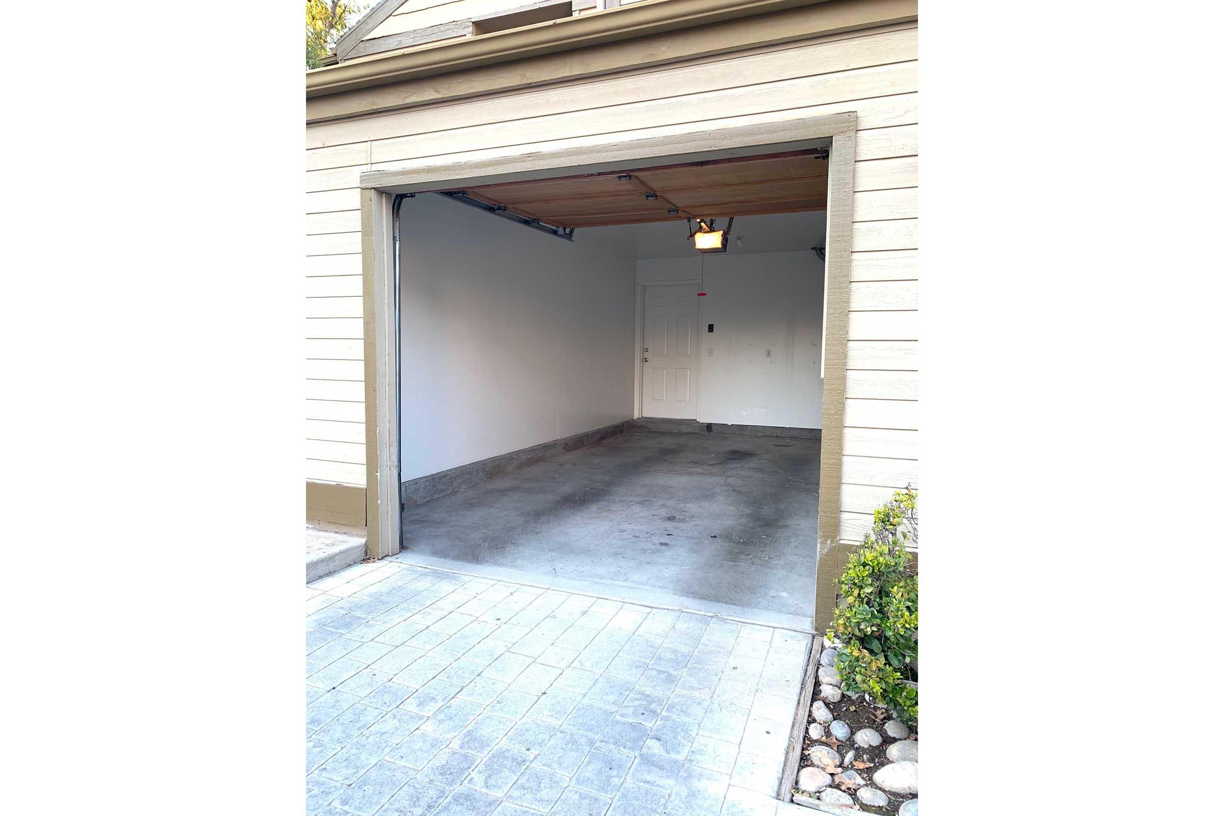 An empty garage with a concrete floor. The garage door is open, revealing a plain interior with no vehicles or items stored. A door on the side wall is visible, and there are light fixtures hanging from the ceiling. The exterior features a paved area and a small garden bed with stones and plants.