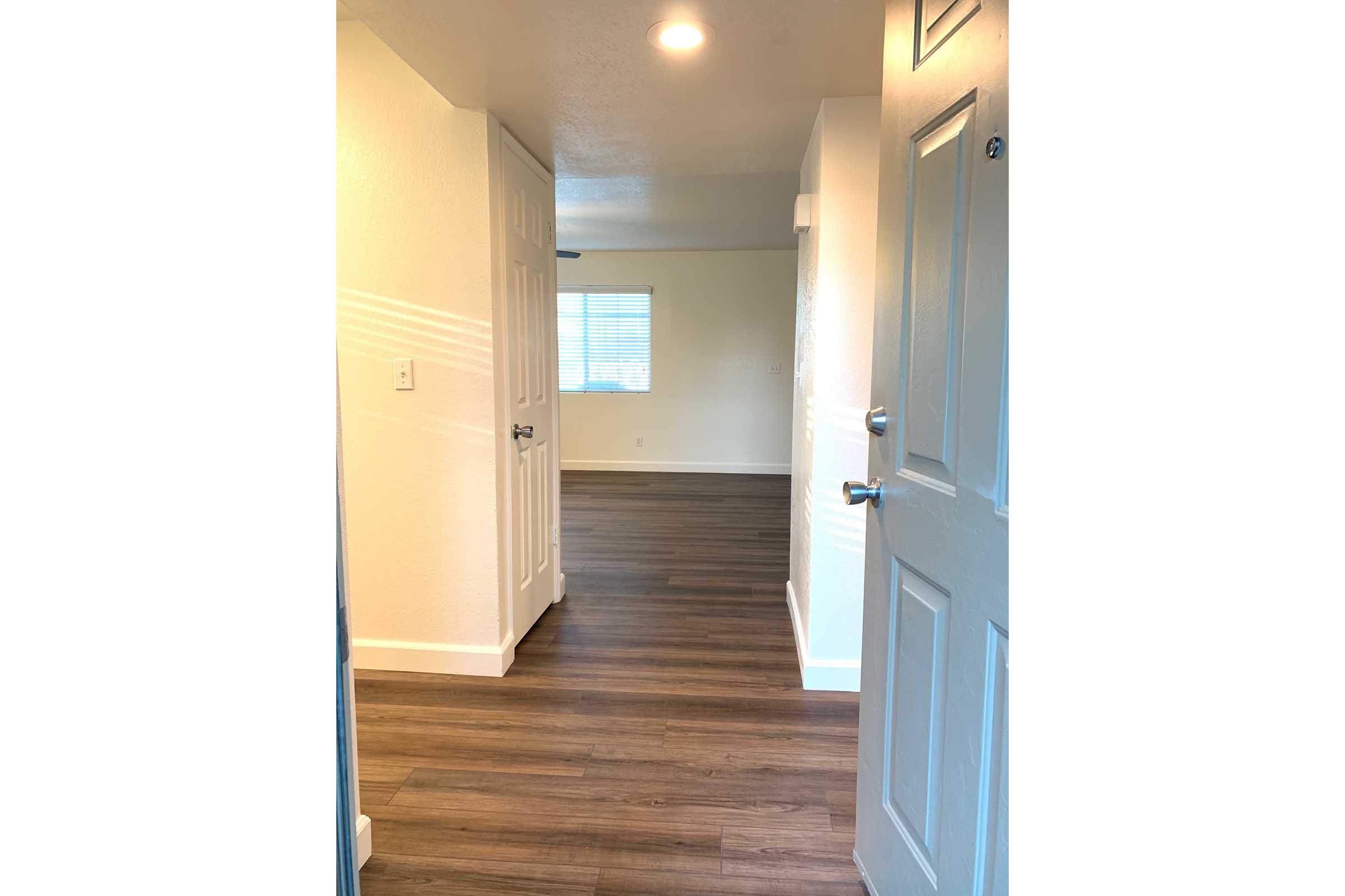 A well-lit entrance hallway leading to a small living area. The flooring is a modern wood-style laminate, and the walls are painted white. There are two doors visible on the left side, and a window at the end of the hallway allows natural light to enter the space.