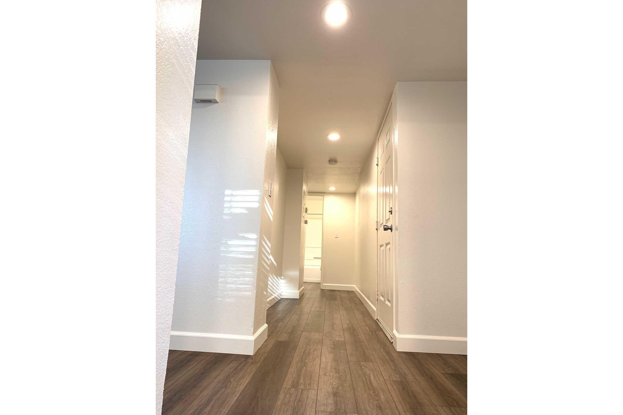 A well-lit hallway featuring light-colored walls and a modern, minimalist design. The floor is covered with wooden planks, and there are several recessed ceiling lights. At the end of the hallway, a door can be seen, leading to another room. The overall atmosphere is clean and inviting.