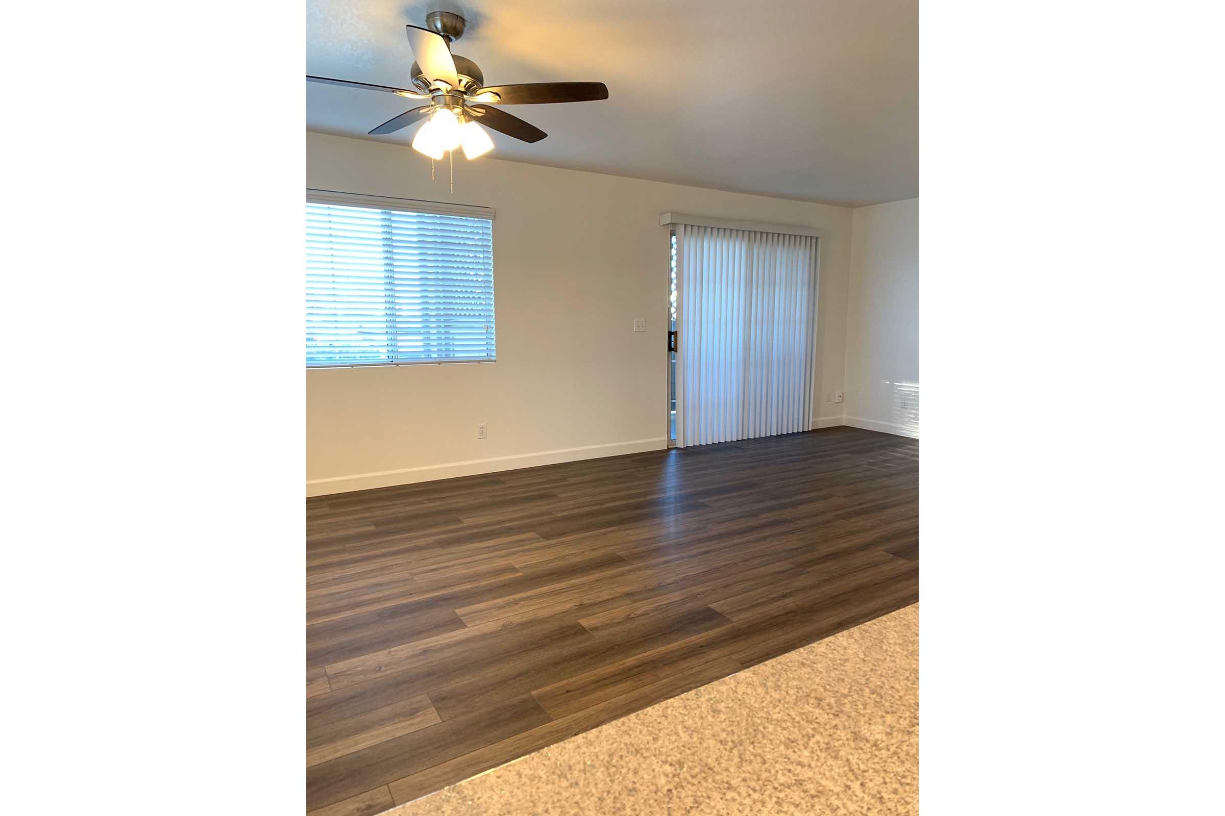 A spacious, well-lit living area featuring a ceiling fan, large window with blinds, and sliding glass door. The flooring is hardwood-style, and the walls are painted a neutral color, creating a bright and inviting atmosphere.