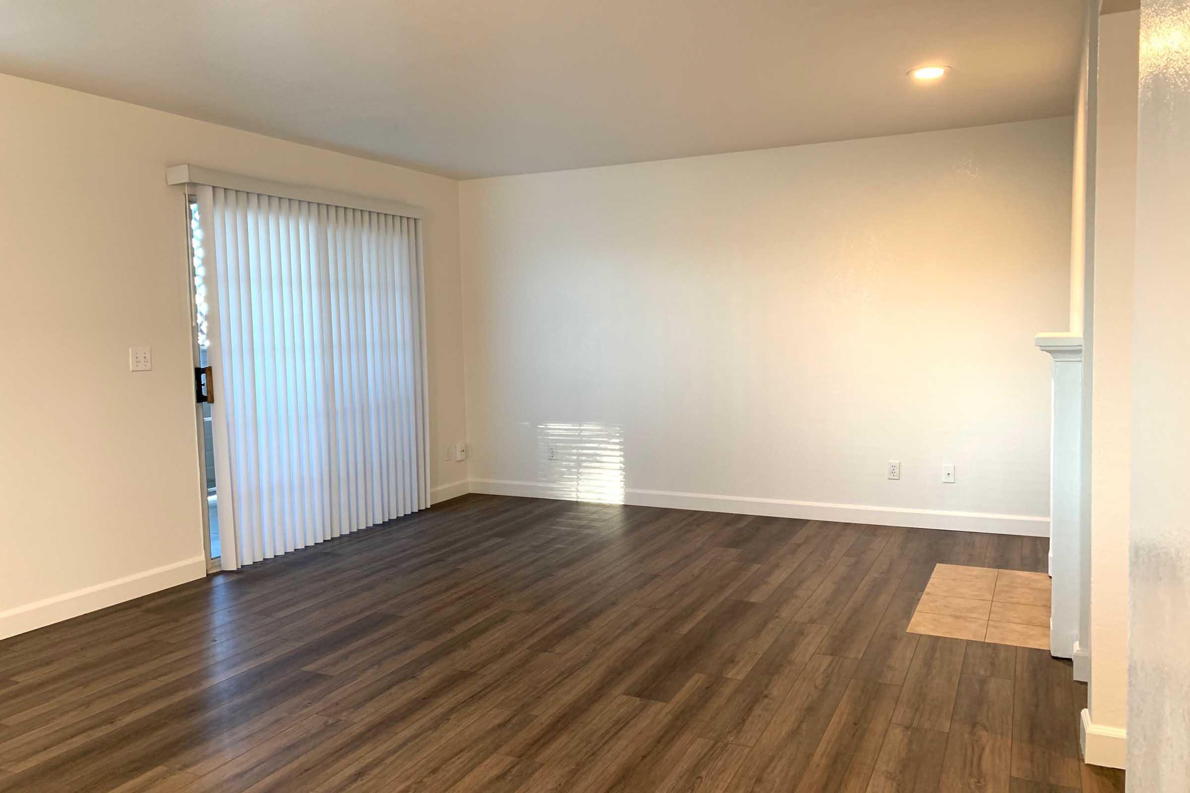 A spacious, empty living room featuring light-colored walls, a hardwood floor, and a sliding glass door with vertical blinds. The room is well-lit and includes a small tiled area on the floor, highlighting an inviting atmosphere ready for furnishings.