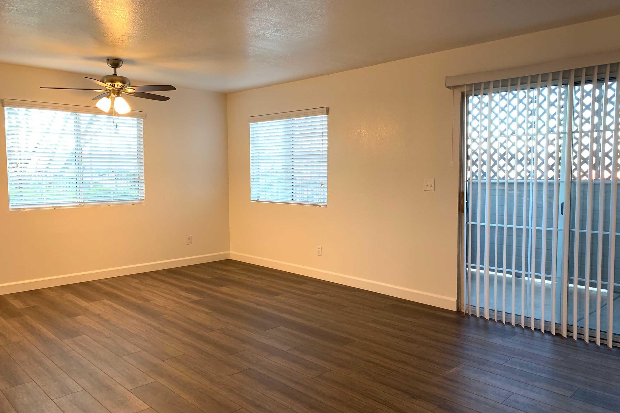 A well-lit empty room with a ceiling fan, two windows with blinds, and sliding glass doors leading to a balcony. The floor is covered with laminate wood, and the walls are painted a light color, creating a clean and spacious atmosphere.