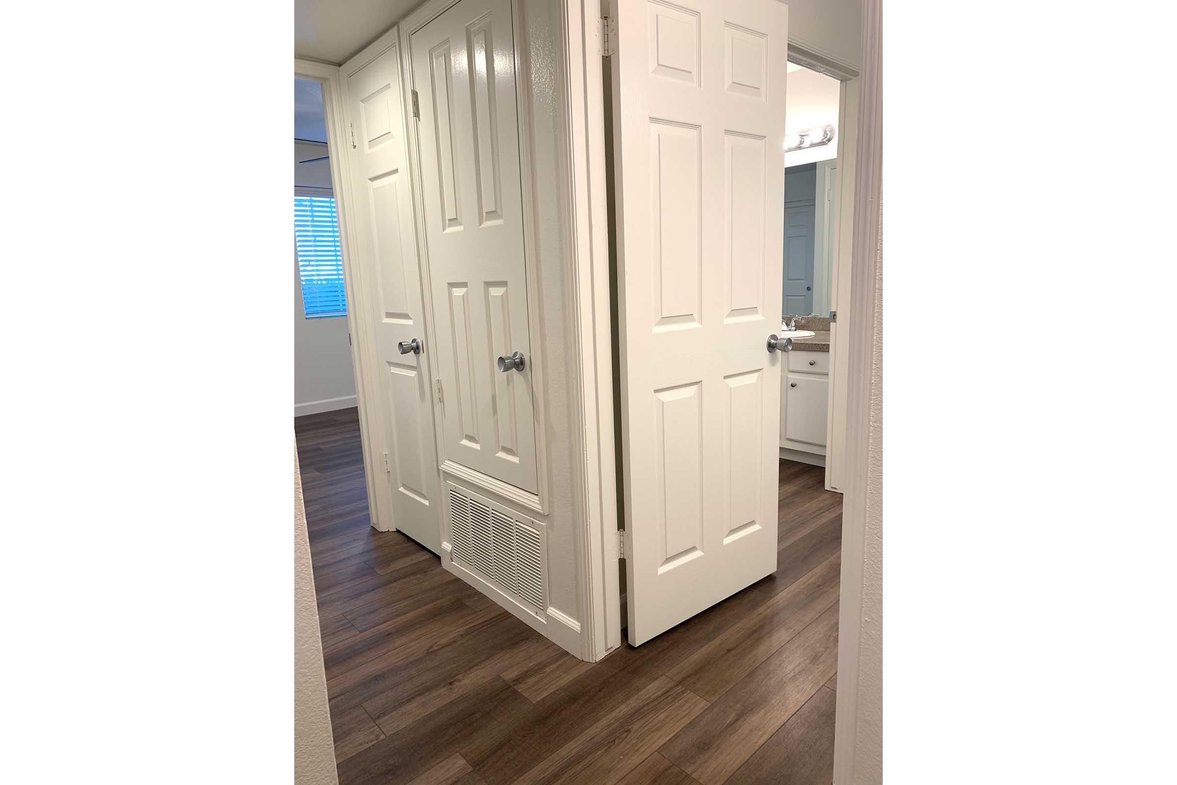 A well-lit hallway featuring two closed white doors on either side, leading to a bathroom area. The floor is covered in wooden planks, and there is a vent on the wall. Natural light is coming from a window at the far end, enhancing the bright and airy feel of the space.