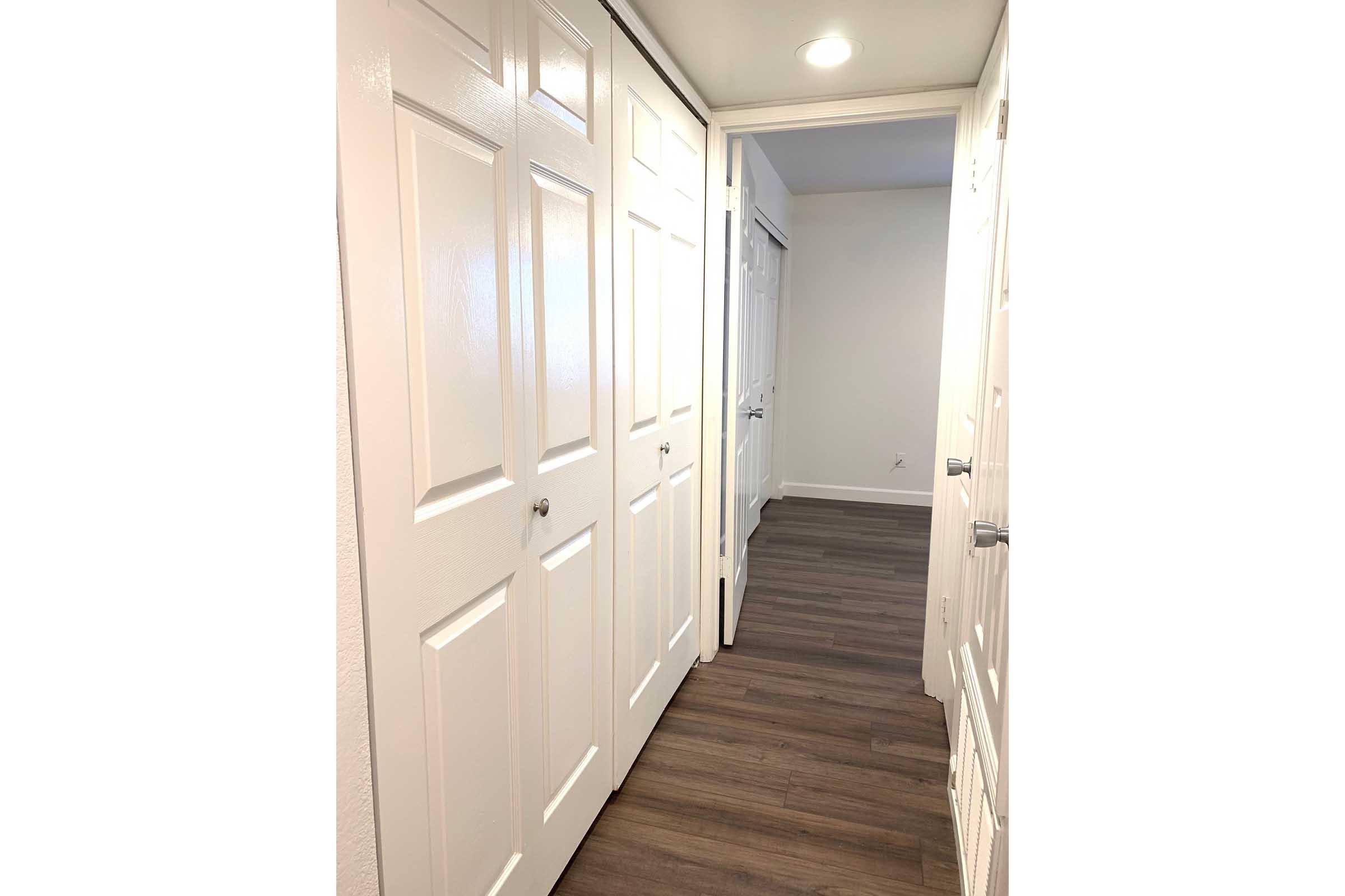 A well-lit hallway featuring multiple white closet doors with modern handles on one side and a continuation of a neutral-colored wall leading to another room. The flooring is a warm wood-tone laminate, contributing to a clean and inviting atmosphere.