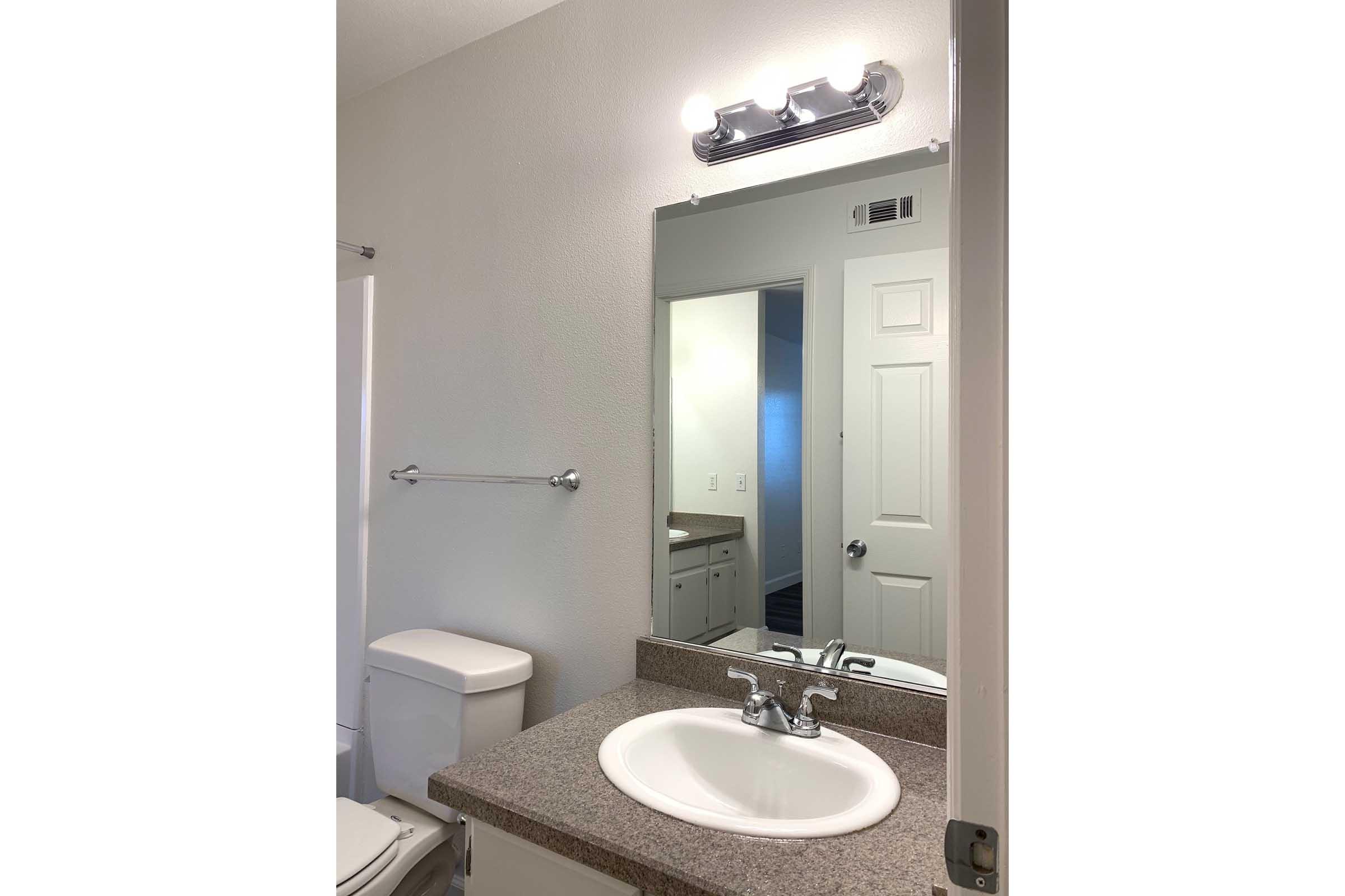 A clean, modern bathroom featuring a white sink with chrome faucet, a large mirror above it, and a light fixture. There's a white toilet in the corner, and a towel bar mounted on the wall. A door leads to another room, reflecting a well-lit and organized space with neutral-colored walls.