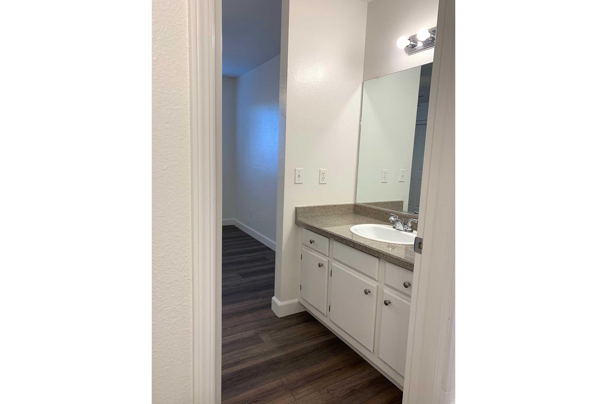 A well-lit bathroom featuring a vanity with a sink and a large mirror. The walls are painted white, and the floor is made of laminate wood. The design is modern with clean lines, and there is a doorway leading to another room in the background.