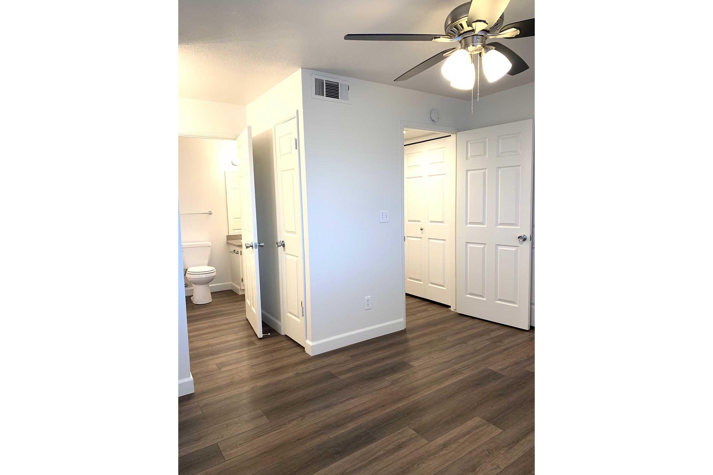Interior view of a well-lit room featuring a ceiling fan, smooth walls, and laminate flooring. There are two doors leading to a bathroom and a closet, with a visible white toilet in the background. The room has a clean, modern appearance, contributing to a spacious feel.