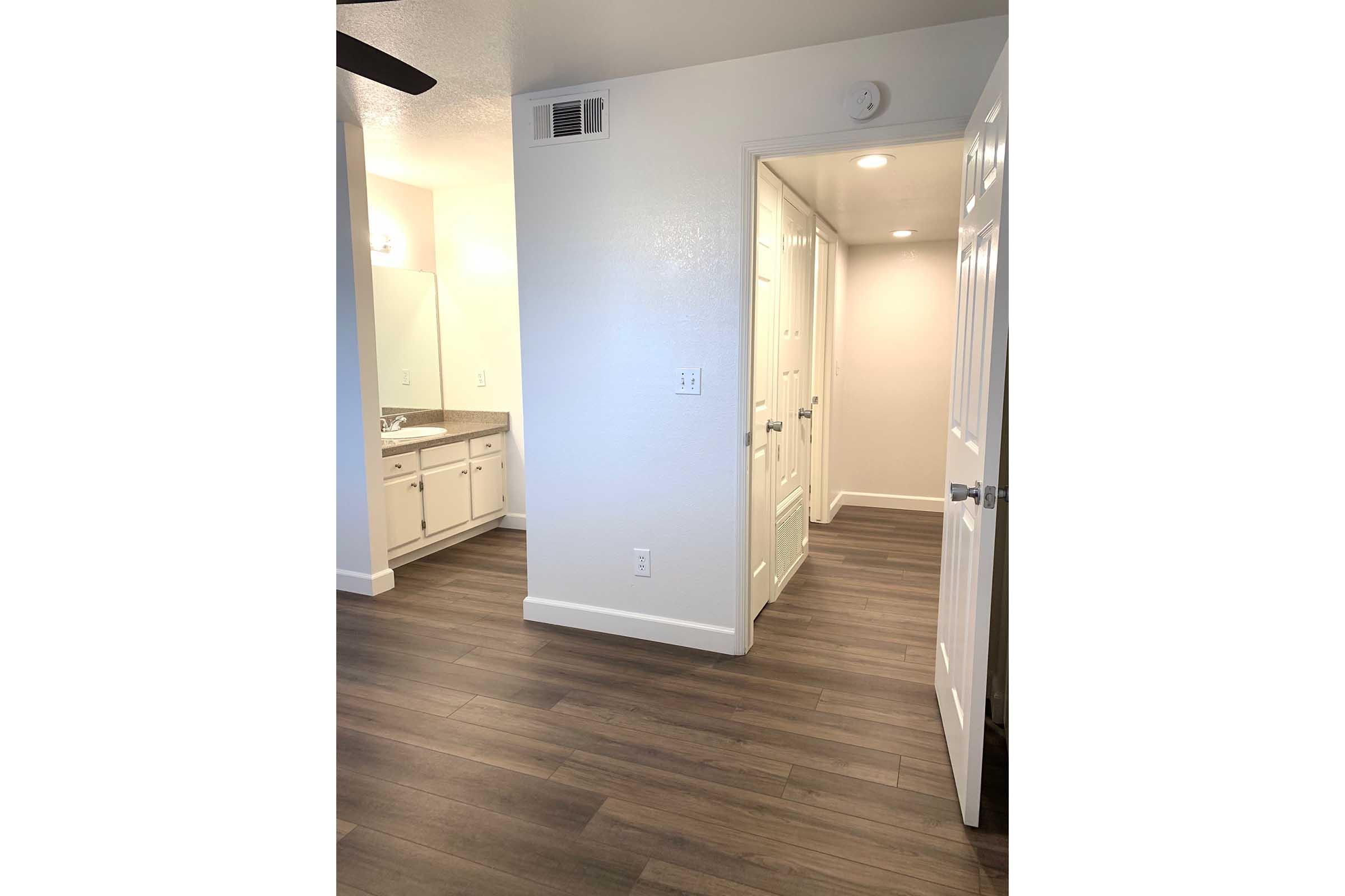 A well-lit interior space featuring a modern bathroom on the left with a large mirror and vanity, and a hallway leading to another room on the right. The floor is made of wooden laminate, and the walls are painted a clean white, creating a bright and inviting atmosphere.