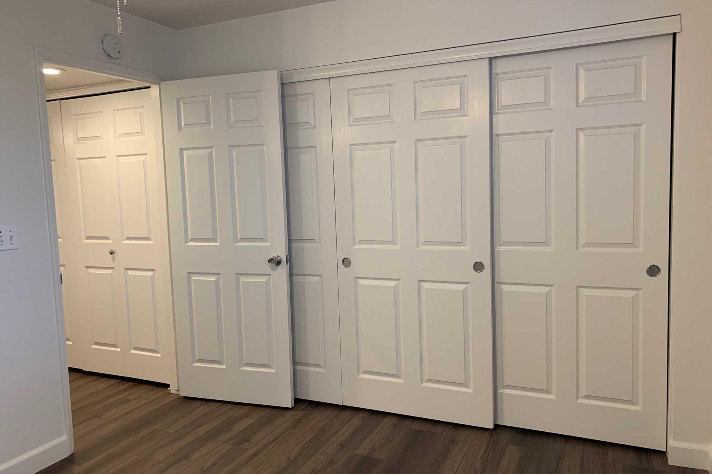 Interior view of a room featuring multiple white doors, including two sliding closet doors and one partially open door leading to another room. The walls are painted light, and the floor is a warm wood-like laminate.