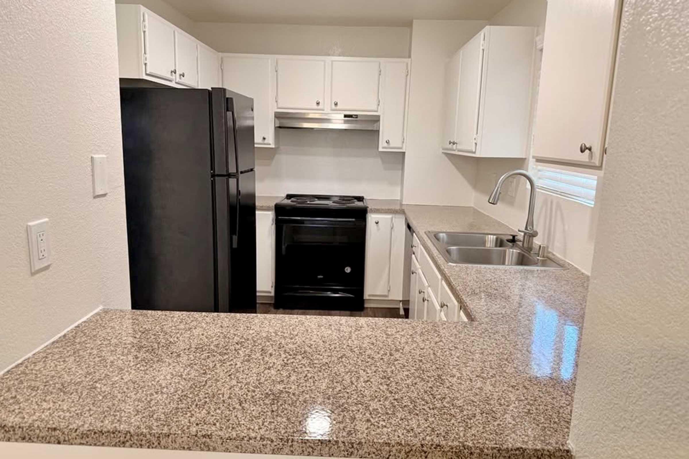 A modern kitchen with white cabinets, black appliances, and a granite countertop. The layout includes a black refrigerator, an oven, and a sink, with natural light coming from a window. The open design connects to a living area, creating a spacious feel.