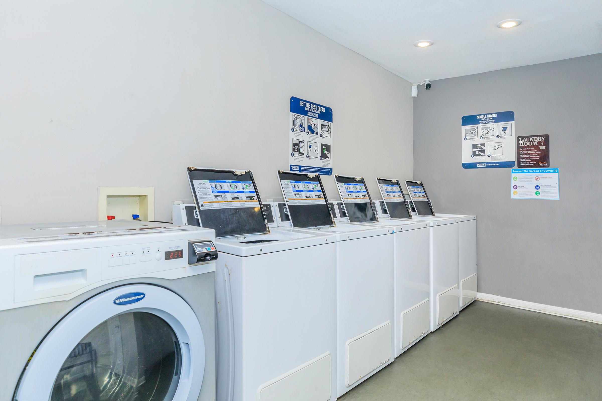 A bright and clean laundry room featuring several stacked washing machines and dryers. The machines are white and well-maintained, lined against a neutral gray wall. Informational signs are mounted above the machines, providing usage instructions and laundry rules. The floor is plain and uncluttered, contributing to a tidy atmosphere.