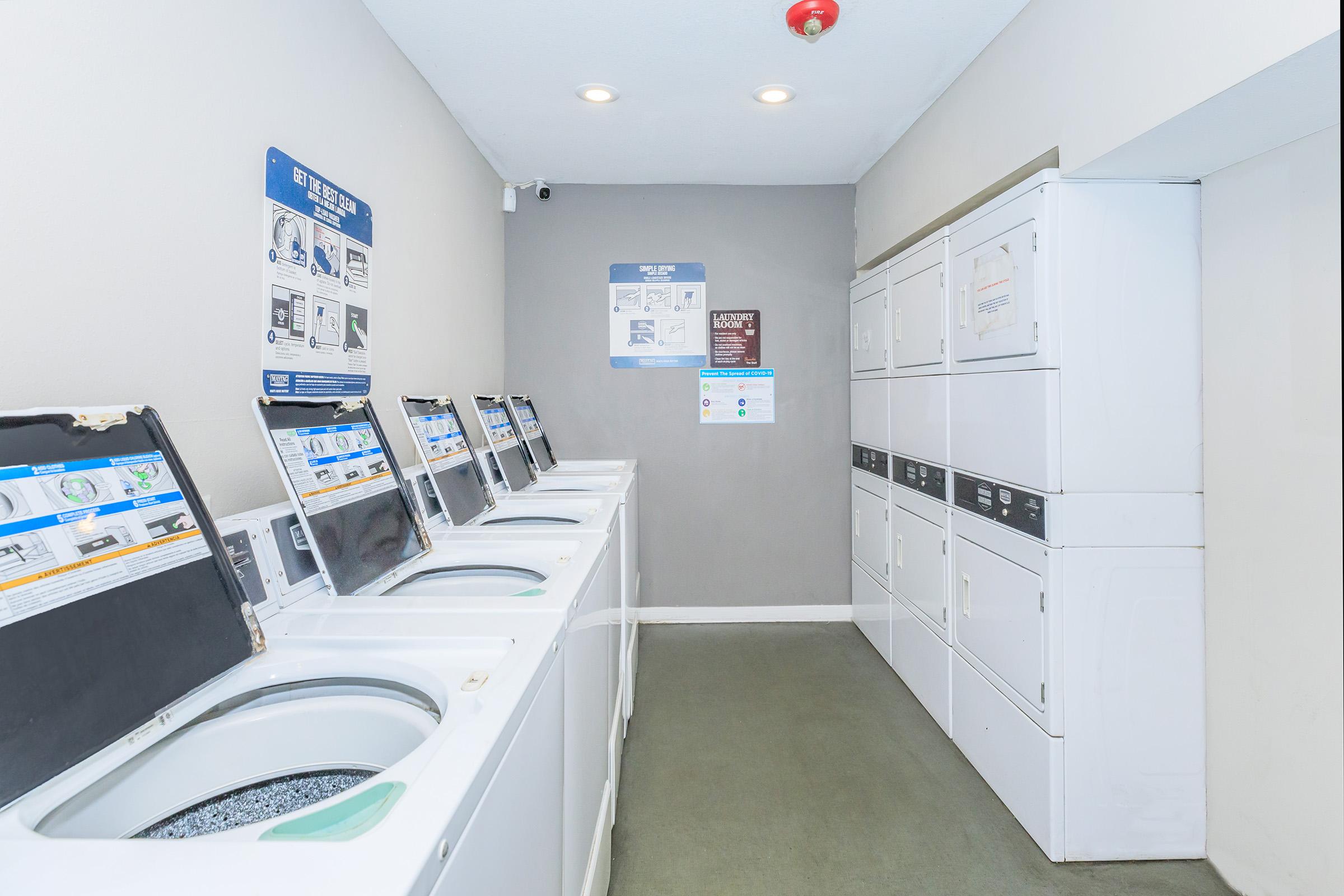 A clean laundry room featuring several white washing machines aligned against a wall. There are informational posters on the wall, and a row of white dryers is visible on the right. The flooring is gray, and the space is well-lit with a ceiling light, contributing to a tidy and organized atmosphere.
