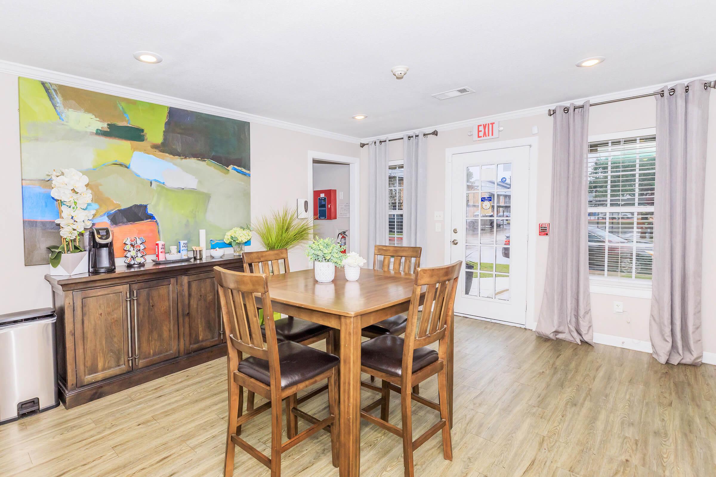 A cozy dining area featuring a wooden table with four chairs, a large abstract painting on the wall, a sideboard with decorative plants, and a coffee station. Natural light streams in through a door with a view of the exterior. Elegant curtains frame the windows, creating a welcoming atmosphere.