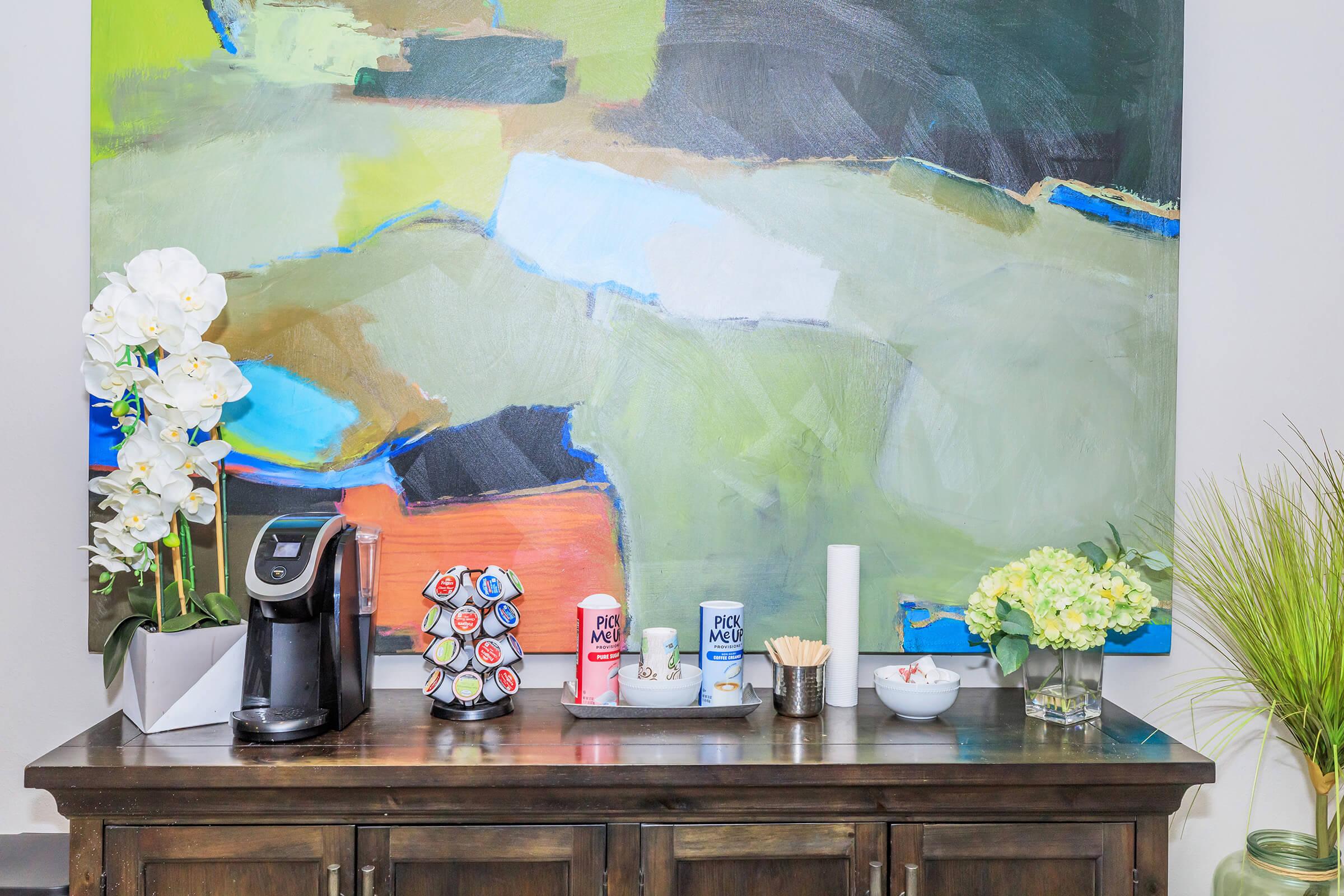 A coffee machine on a wooden table next to a colorful abstract painting. There are coffee pods displayed in a holder, sugar and cream containers, a small bowl with snacks, and decorative flowers in a vase, creating a welcoming coffee station.