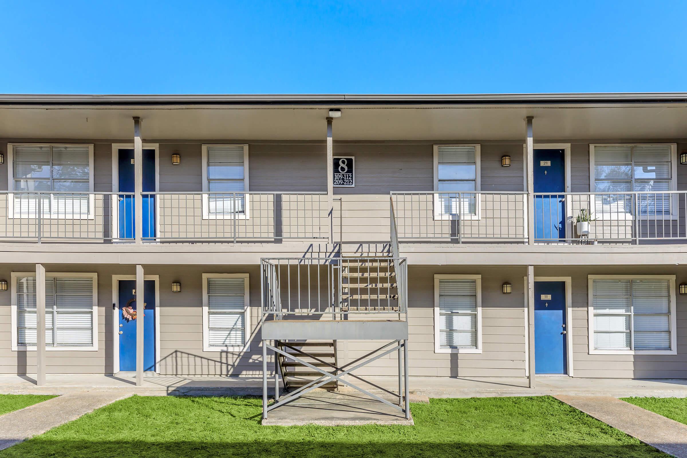 Two-story apartment building with a light gray exterior. There are four units visible, each with a blue door and white window shutters. A central staircase leads to the second floor, and a small green lawn is adjacent to the building under a clear blue sky.