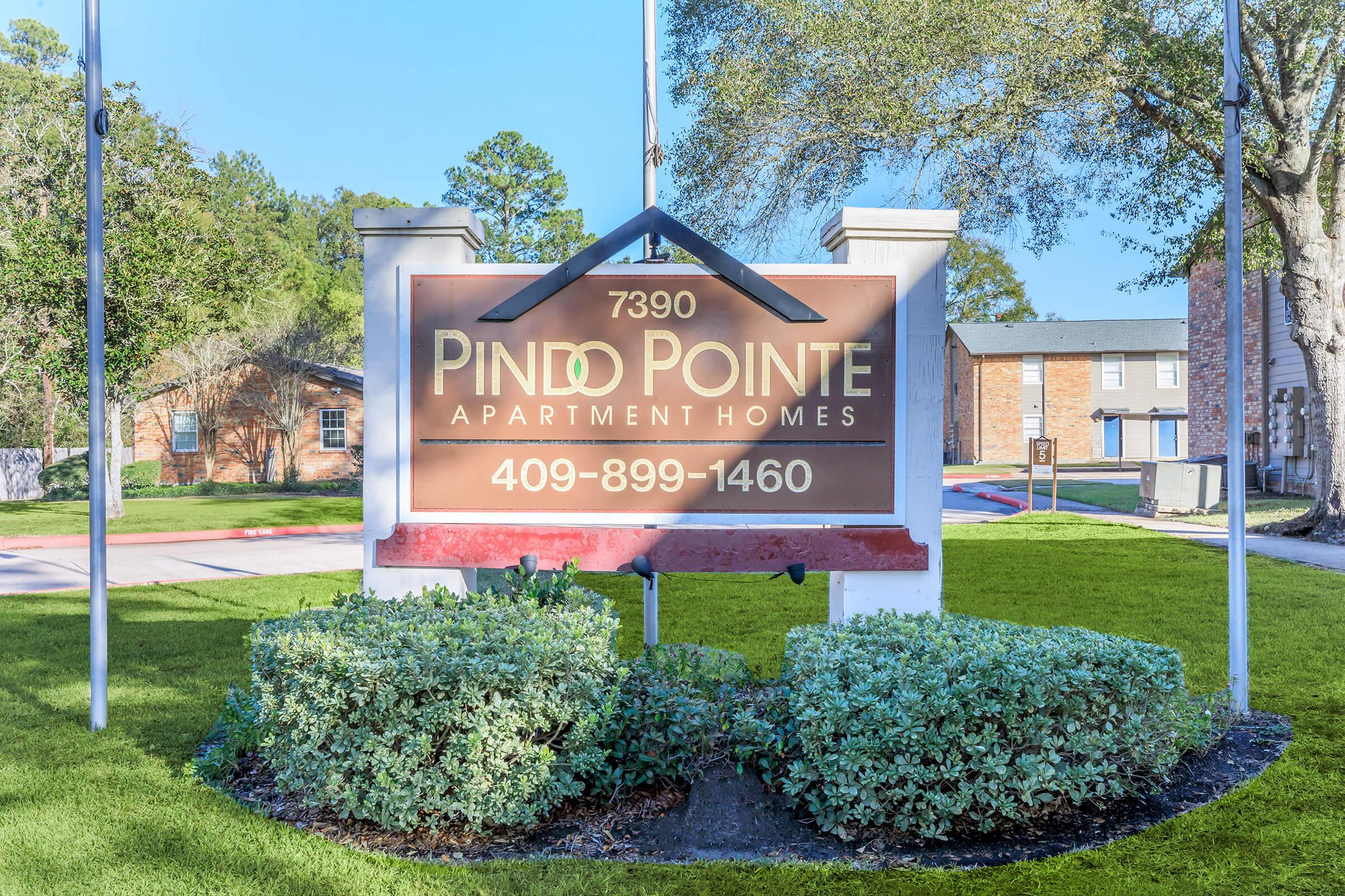 Sign for Pinle Pointe Apartment Homes located at 7390. The sign features a decorative design and contact number 409-899-1460. Surrounding the sign are well-maintained shrubs and grass, with buildings visible in the background under clear skies.