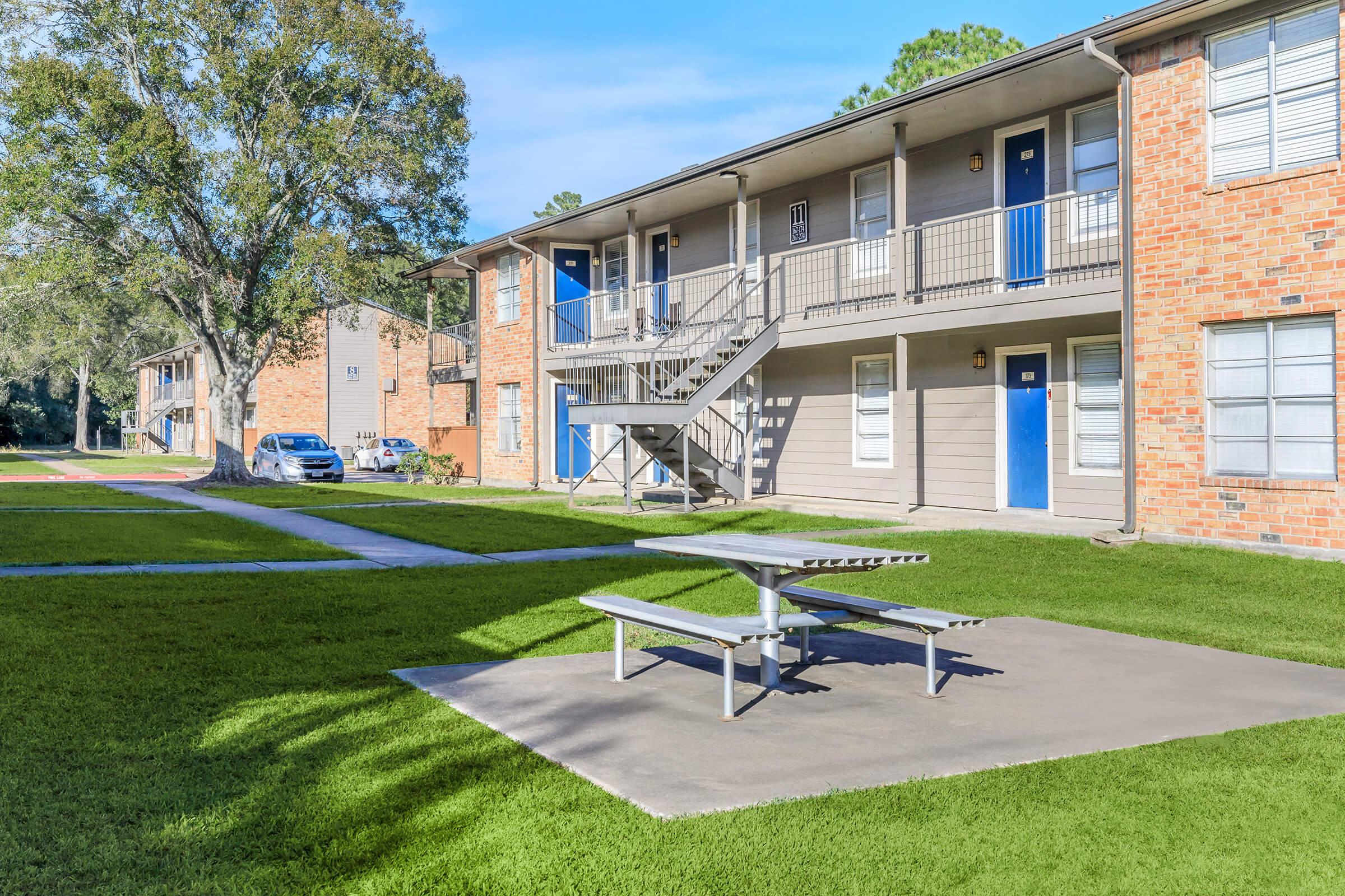 A sunny day at an apartment complex featuring brick buildings with blue doors, surrounded by green lawns. A picnic table is placed on a concrete slab in the center, with a lined pathway leading to the units. A tree offers some shade in the background.