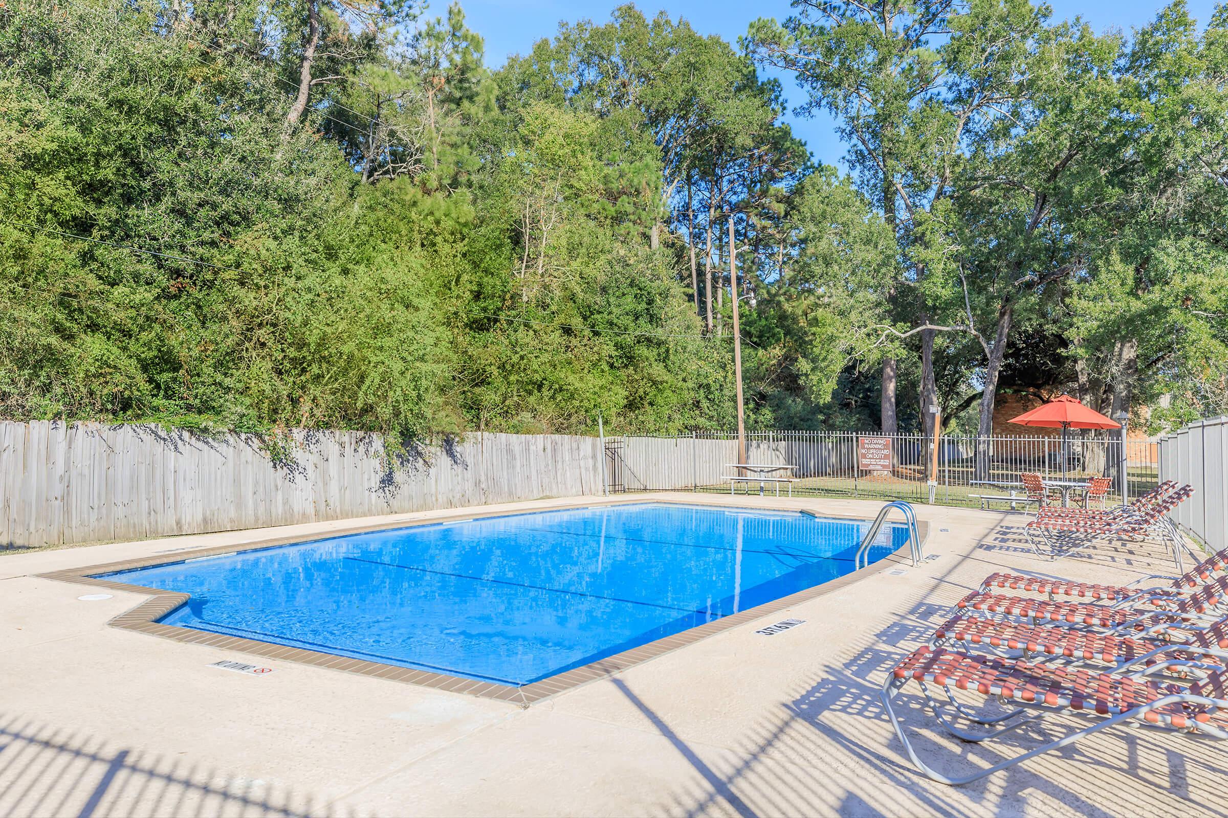 A bright and clear swimming pool surrounded by a concrete deck. Several lounge chairs are lined up beside the pool, and a shaded area with picnic tables is visible in the background. Lush green trees and a wooden fence surround the pool area, creating a serene outdoor setting.