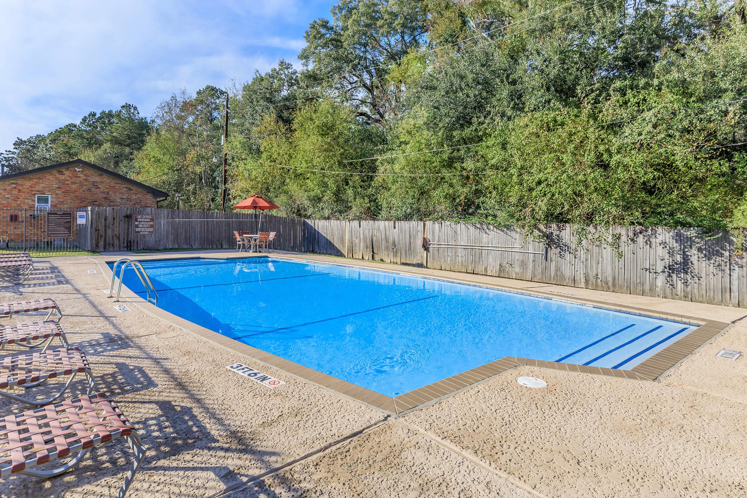 A large outdoor swimming pool surrounded by a concrete deck. There are several lounge chairs arranged around the pool, and a small umbrella is visible in the background. The area is bordered by a wooden fence and lush greenery, with a clear blue sky overhead.