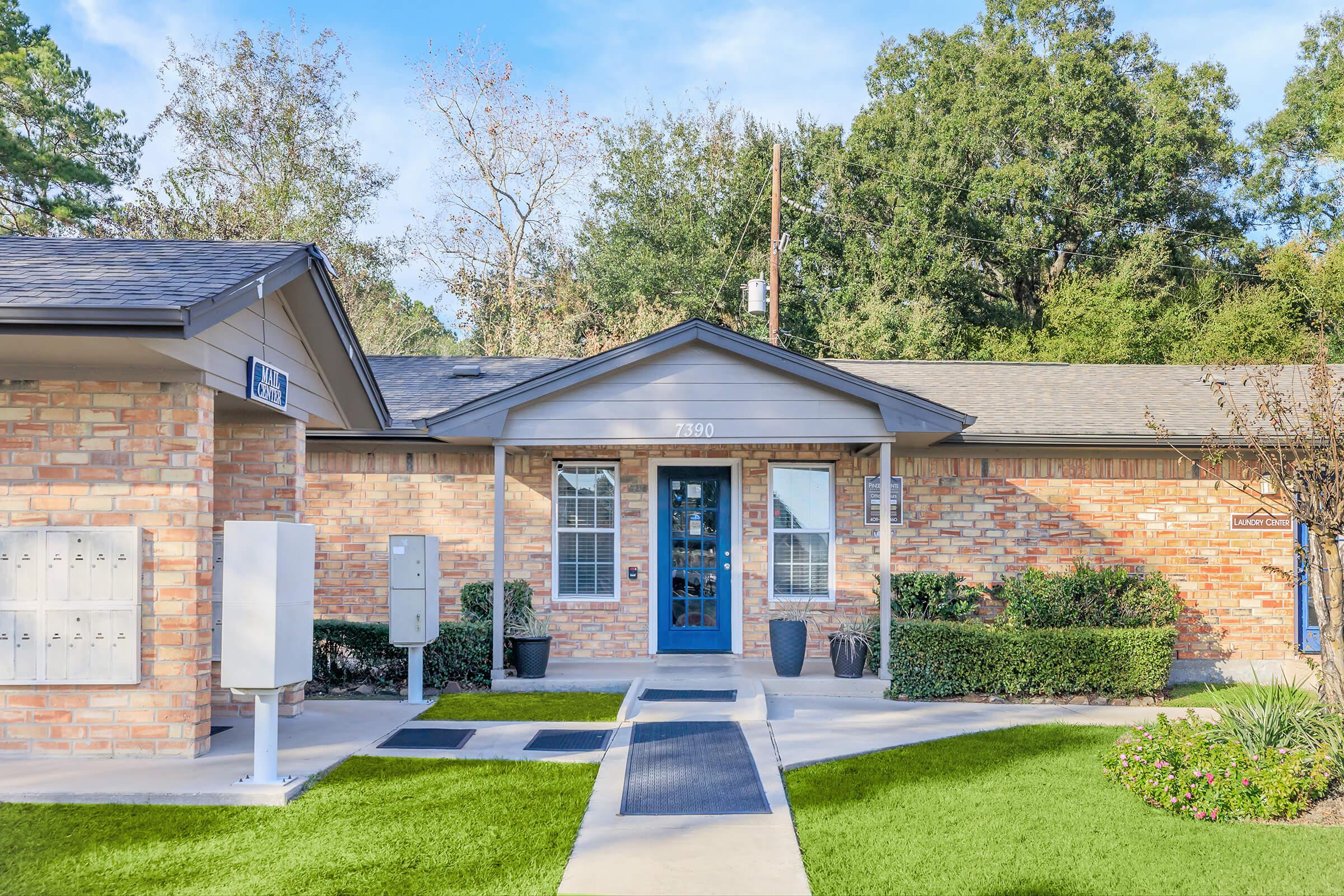 Front entrance of a brick building with a blue door. The pathway is bordered by green grass and small shrubs. Mailboxes are visible to the left, and there are potted plants near the entrance. Trees provide a natural backdrop on a clear day.