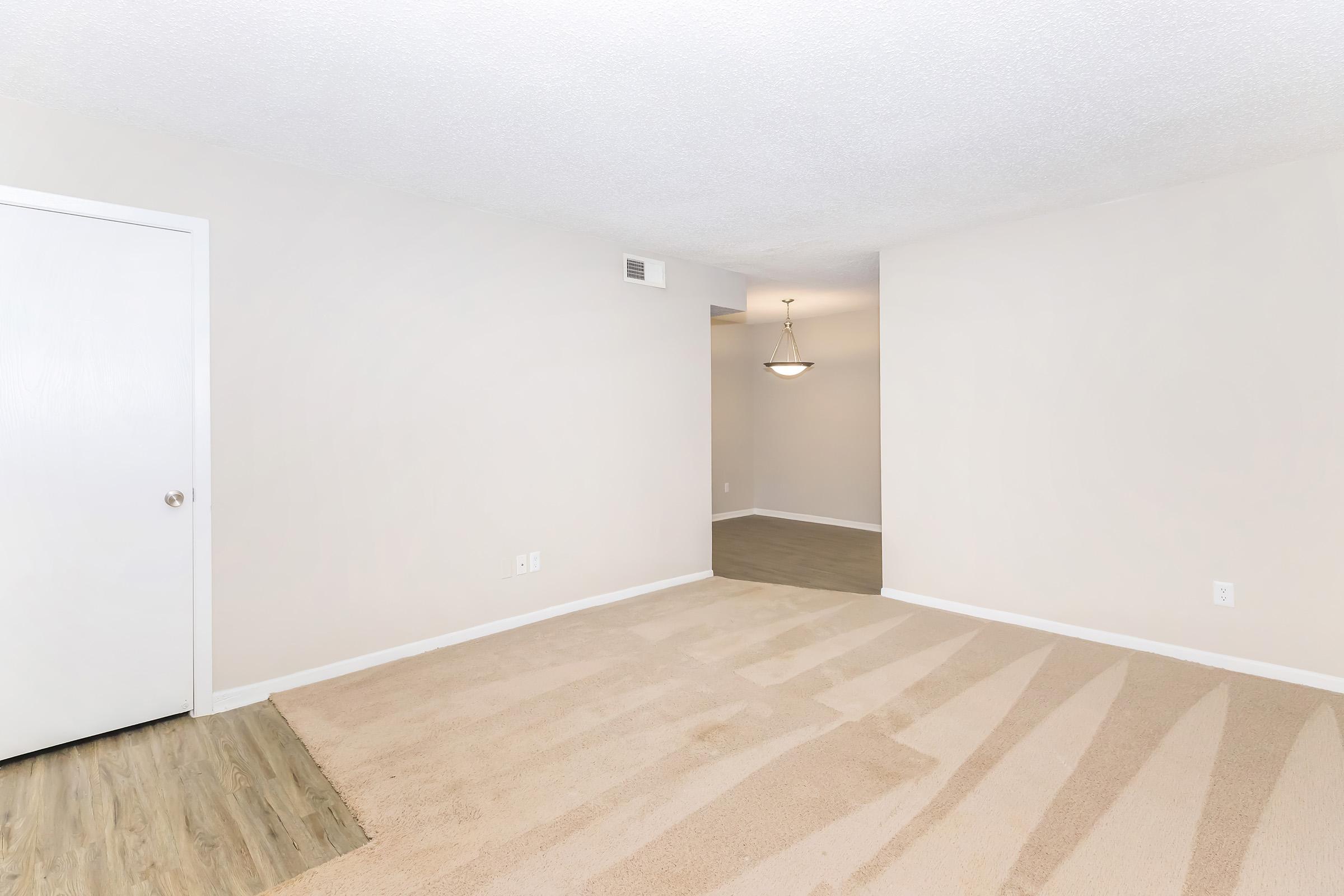 A spacious, empty living room with light beige walls and carpet. A closed white door is on the left side, and a glimpse of a dining area with a light fixture is visible in the background. The floor is a mix of carpet and light wood, creating a warm and inviting atmosphere.