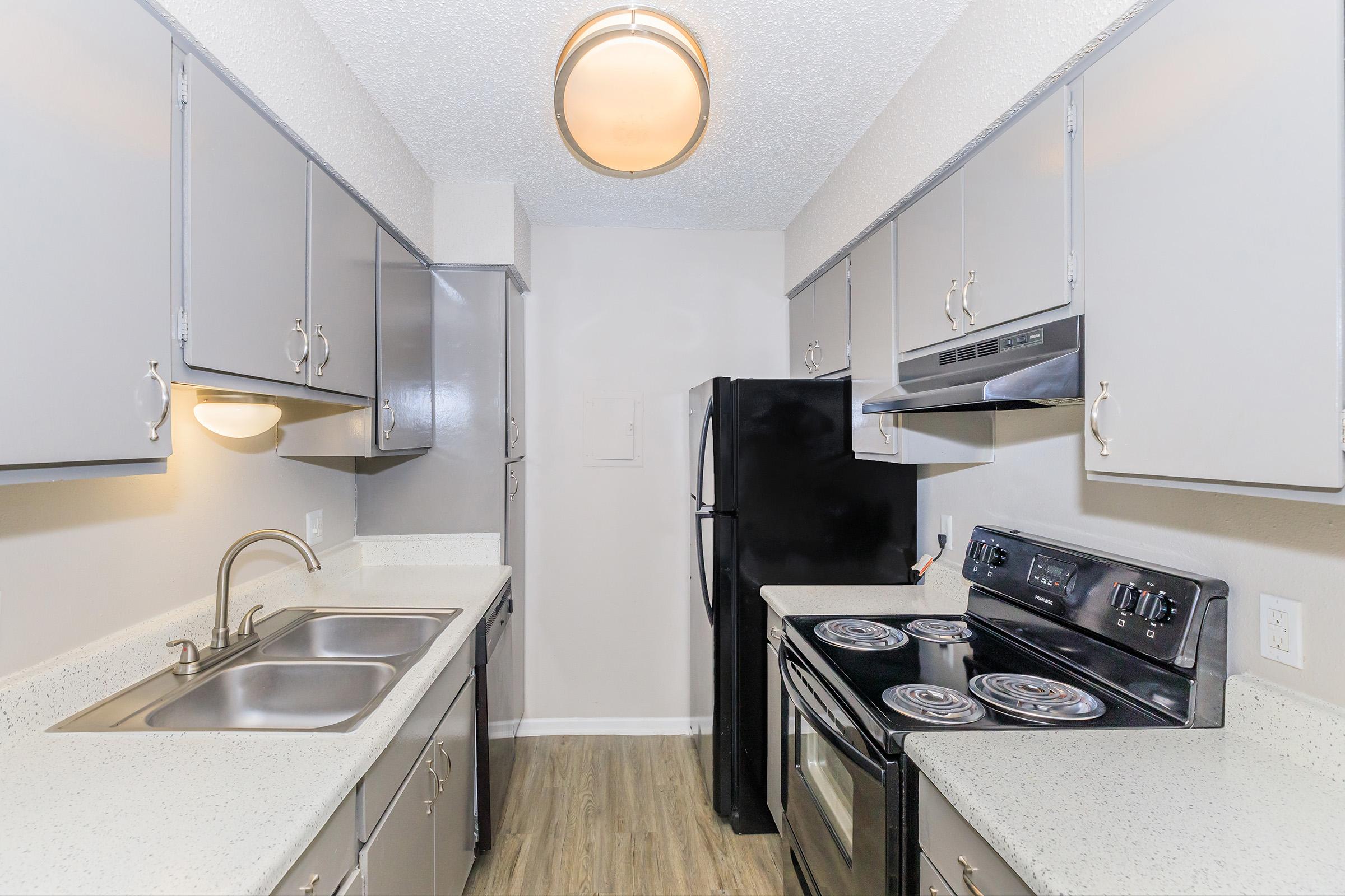 A modern kitchen featuring gray cabinetry, a double sink, an electric stove and oven, and a black refrigerator. The space has a light-colored countertop and laminate flooring, with overhead lighting illuminating the area.