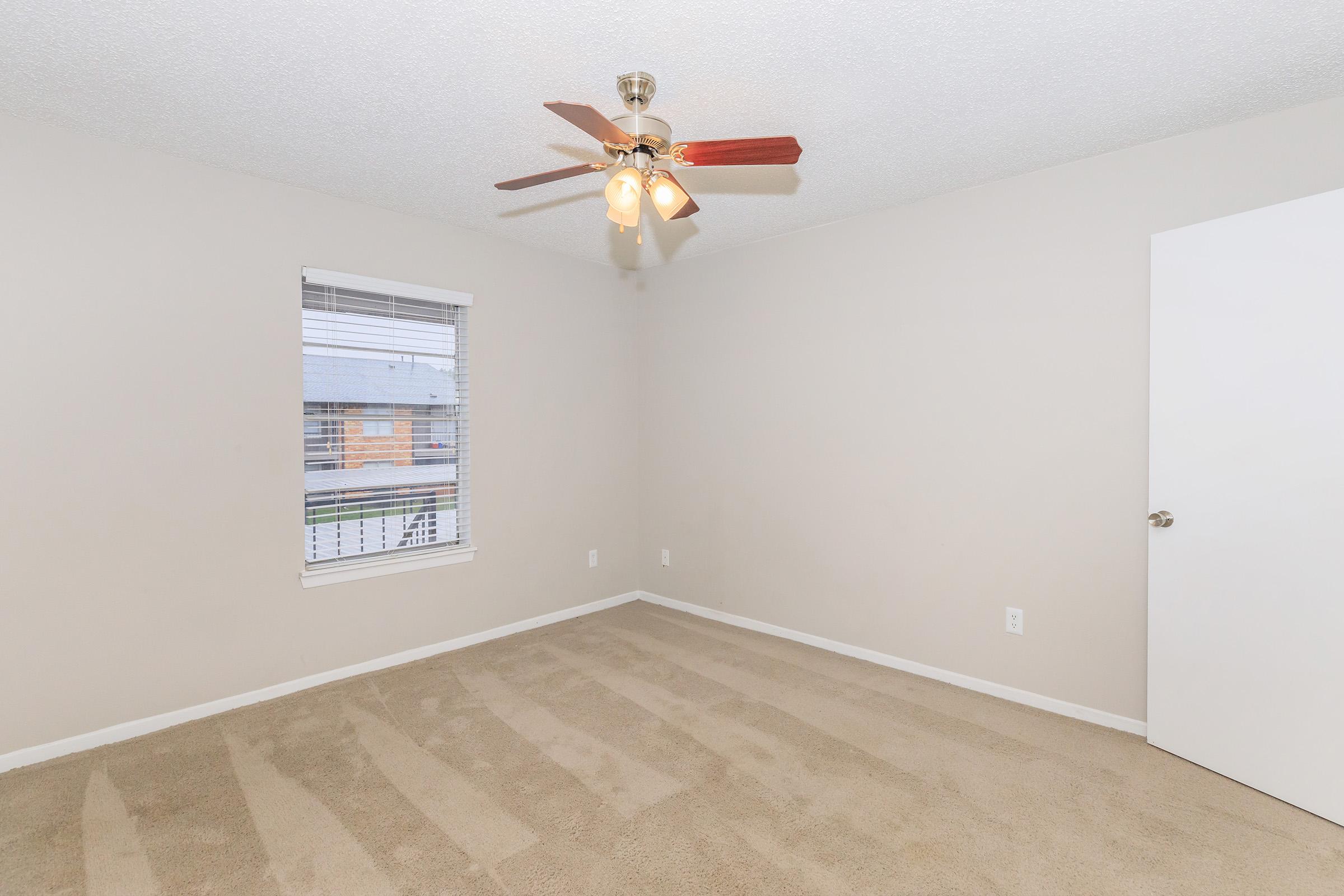 Empty beige bedroom with light tan carpet, a ceiling fan with four lights, and a window with white blinds. A plain white door is visible on the right. The walls are unadorned, creating a simple and clean look.