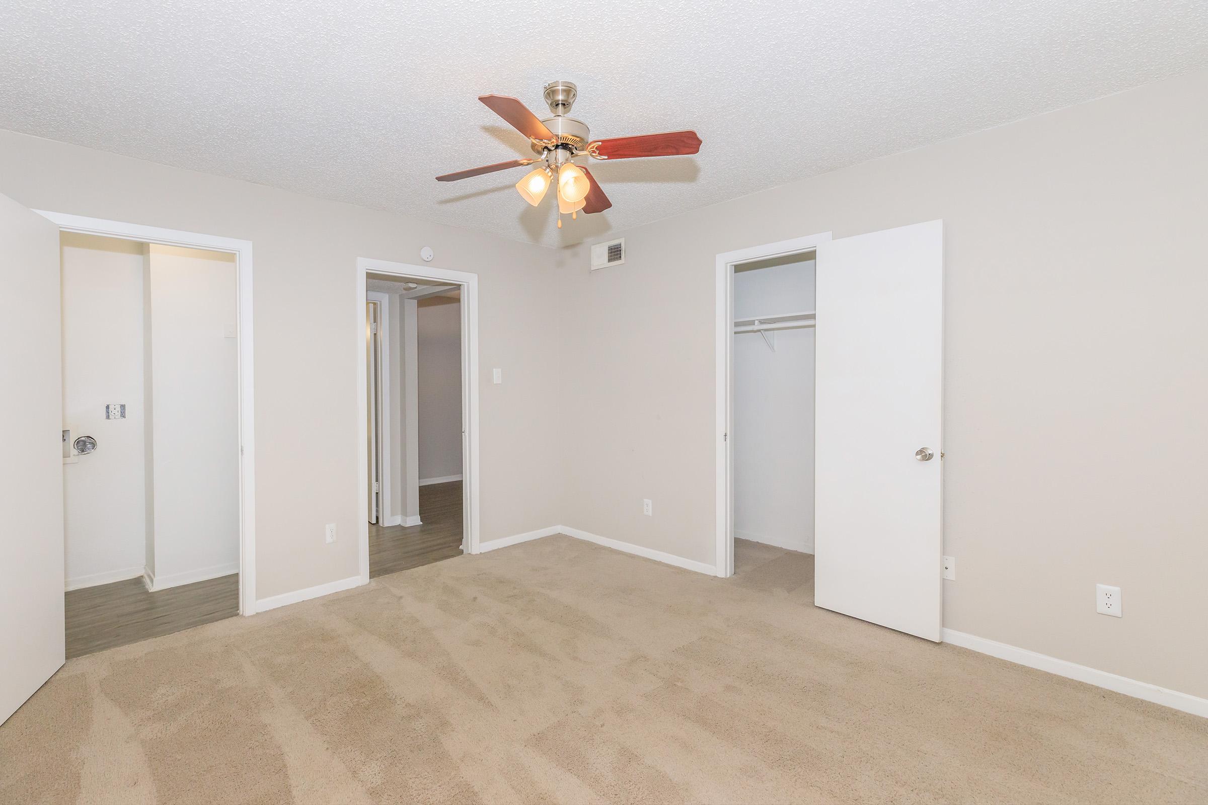 A well-lit, empty room with beige carpet and neutral-colored walls. There are two doors on the left side leading to other spaces and one door on the right, revealing a closet. A ceiling fan with five lights hangs in the center, adding a touch of brightness to the space.