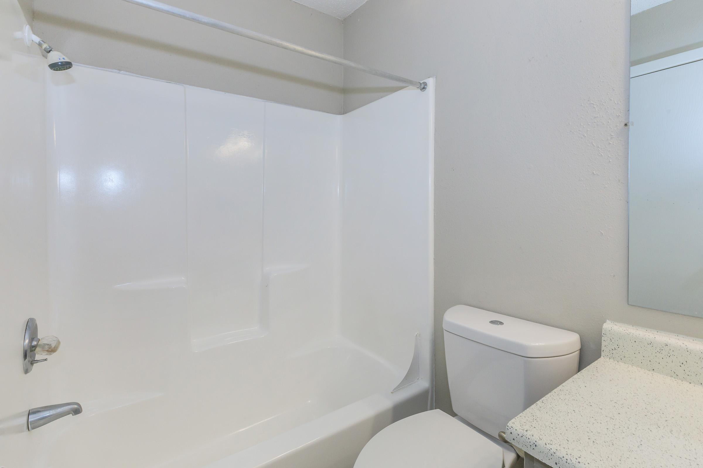 A clean bathroom featuring a white bathtub with a shower curtain, a toilet, and a light-colored countertop. The walls are painted a neutral gray, and a mirror is partially visible above the sink area. The space appears well-maintained and simple in design.