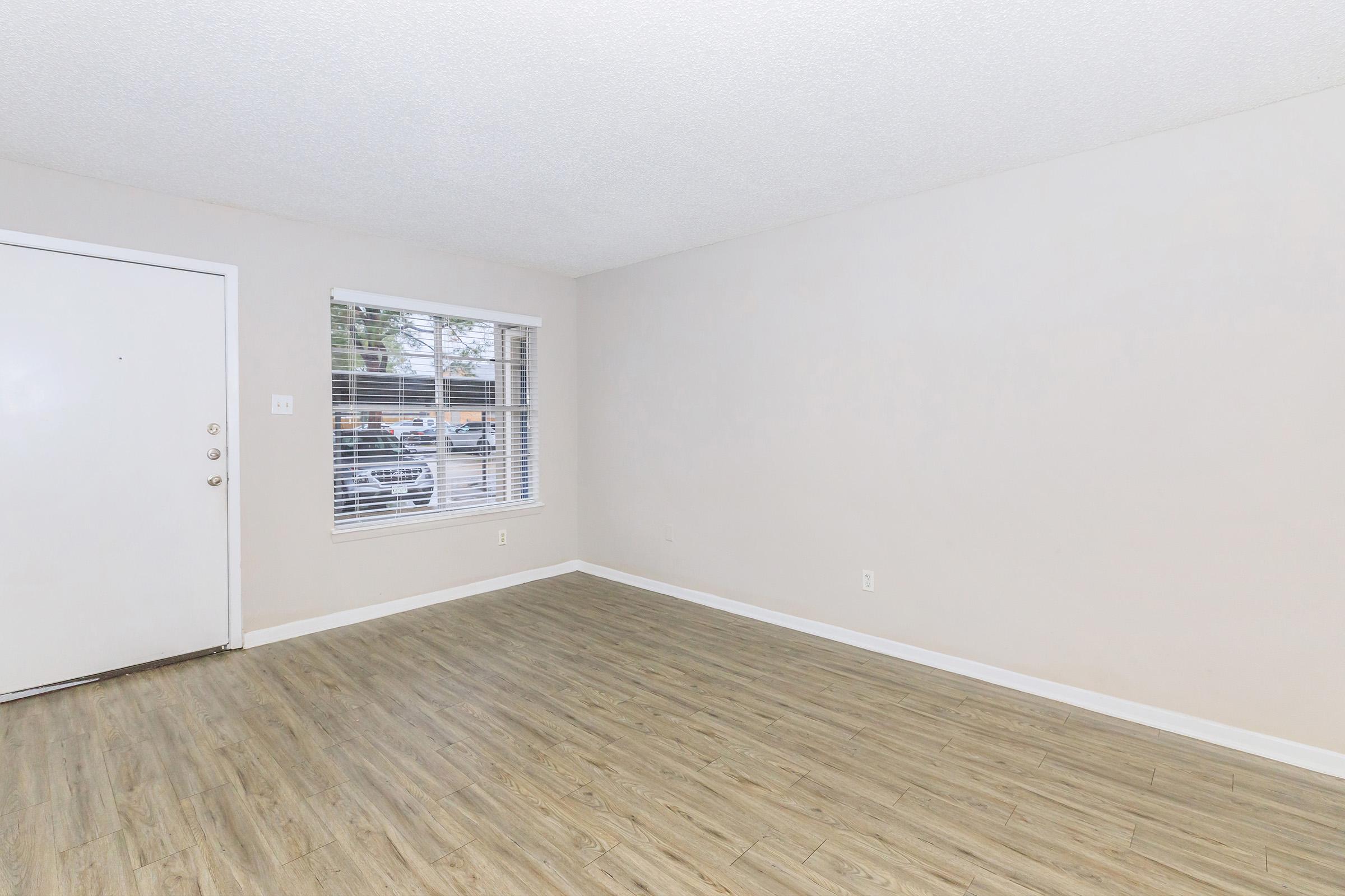 Empty room with light-colored walls and a light wood floor. There is a white door on the left and a window with blinds on the right, letting in natural light. The space is clear of furniture, creating a minimalist and open atmosphere.