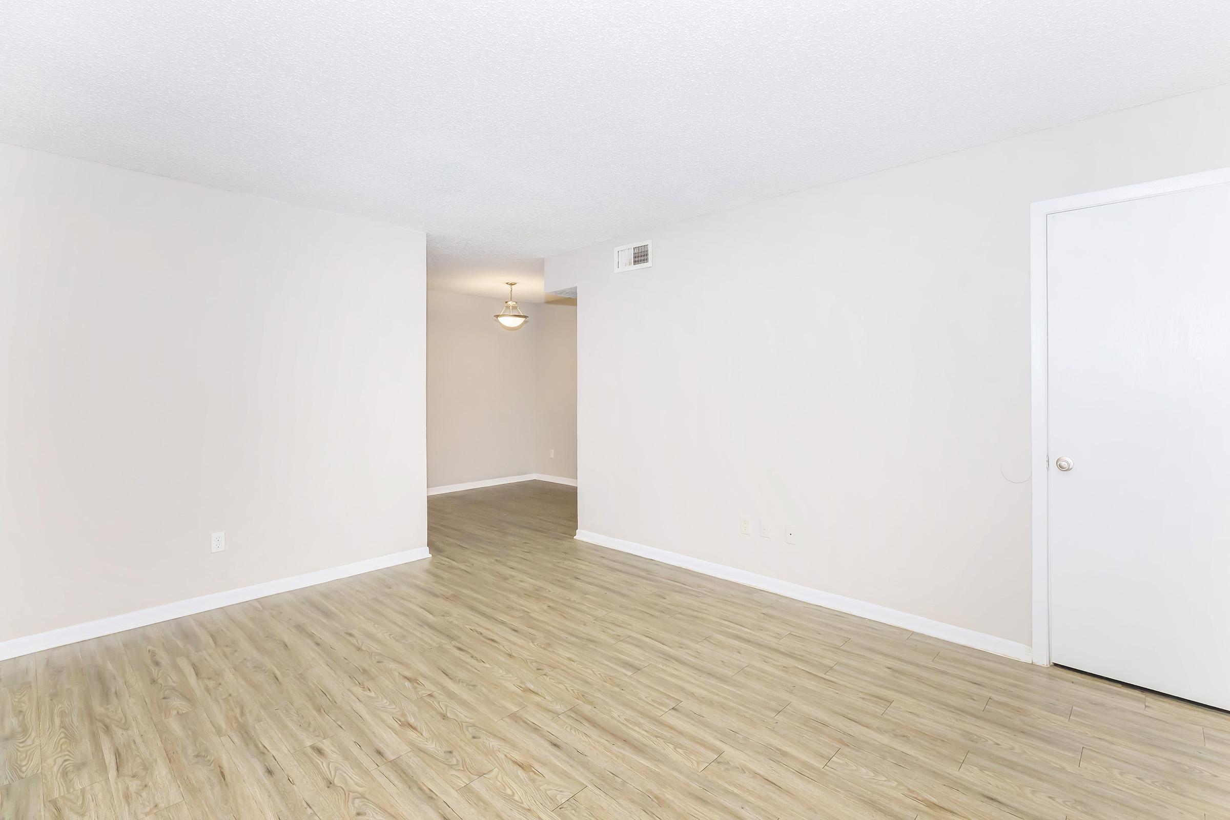 Empty, well-lit room with light-colored walls and a wood-like floor. A doorway is visible on the right, leading to another room. A ceiling light fixture is present. The overall aesthetic is clean and modern, suitable for a living space or apartment.