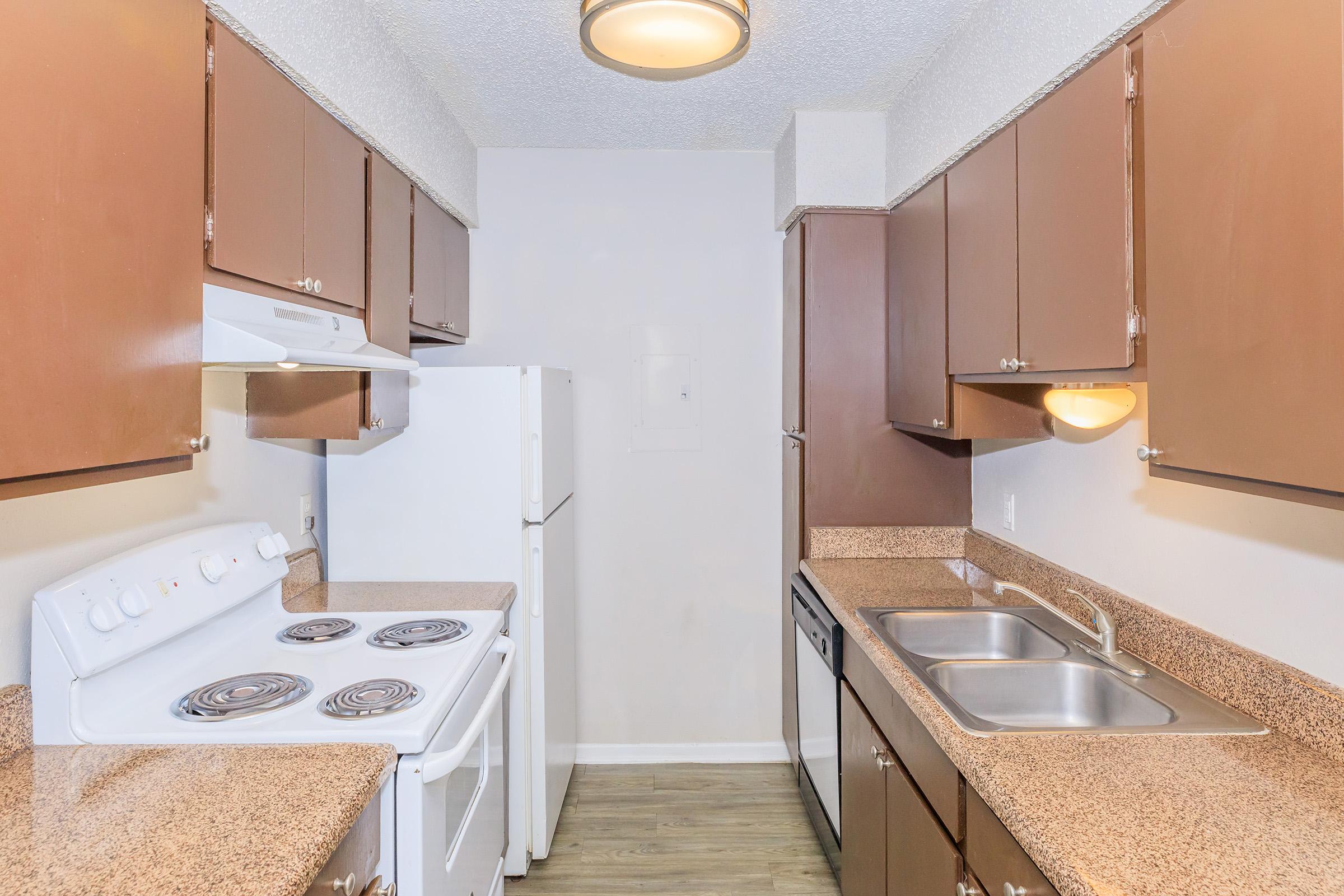A modern kitchen with dark wood cabinets, beige countertops, a white stove, and a white refrigerator. The space includes a double sink, a dishwasher, and overhead lighting, creating a bright and functional cooking area.