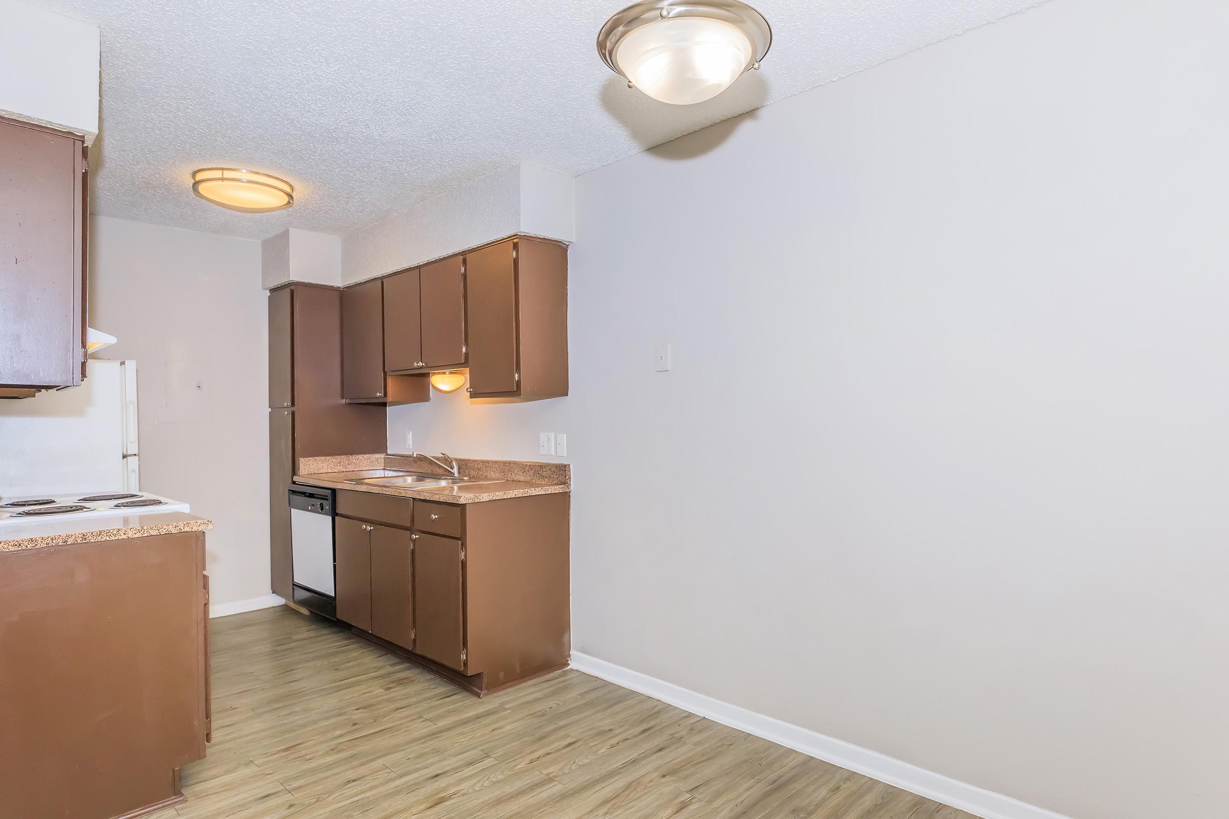 A kitchen with brown cabinets and countertops, a white refrigerator, and a stove. The walls are light-colored, and there is a light fixture on the ceiling. The floor is a light wood laminate, creating a modern and clean appearance. The space is brightly lit and uncluttered.