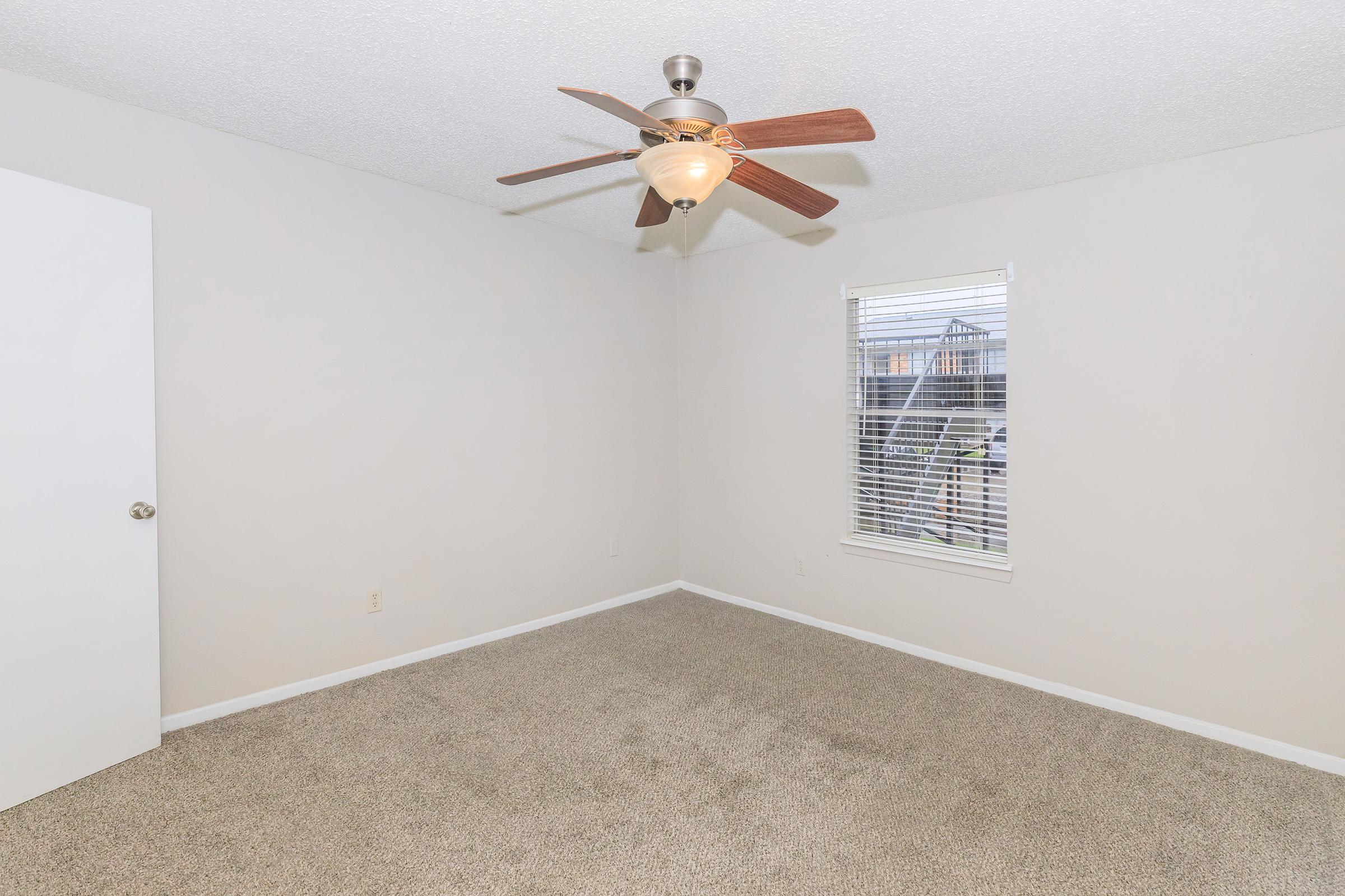 A spacious, neutral-colored bedroom featuring a ceiling fan, a window with blinds, and light-colored carpet. One wall has a door leading to another space, adding depth to the room's layout. The overall ambiance is bright and inviting, perfect for personalization.