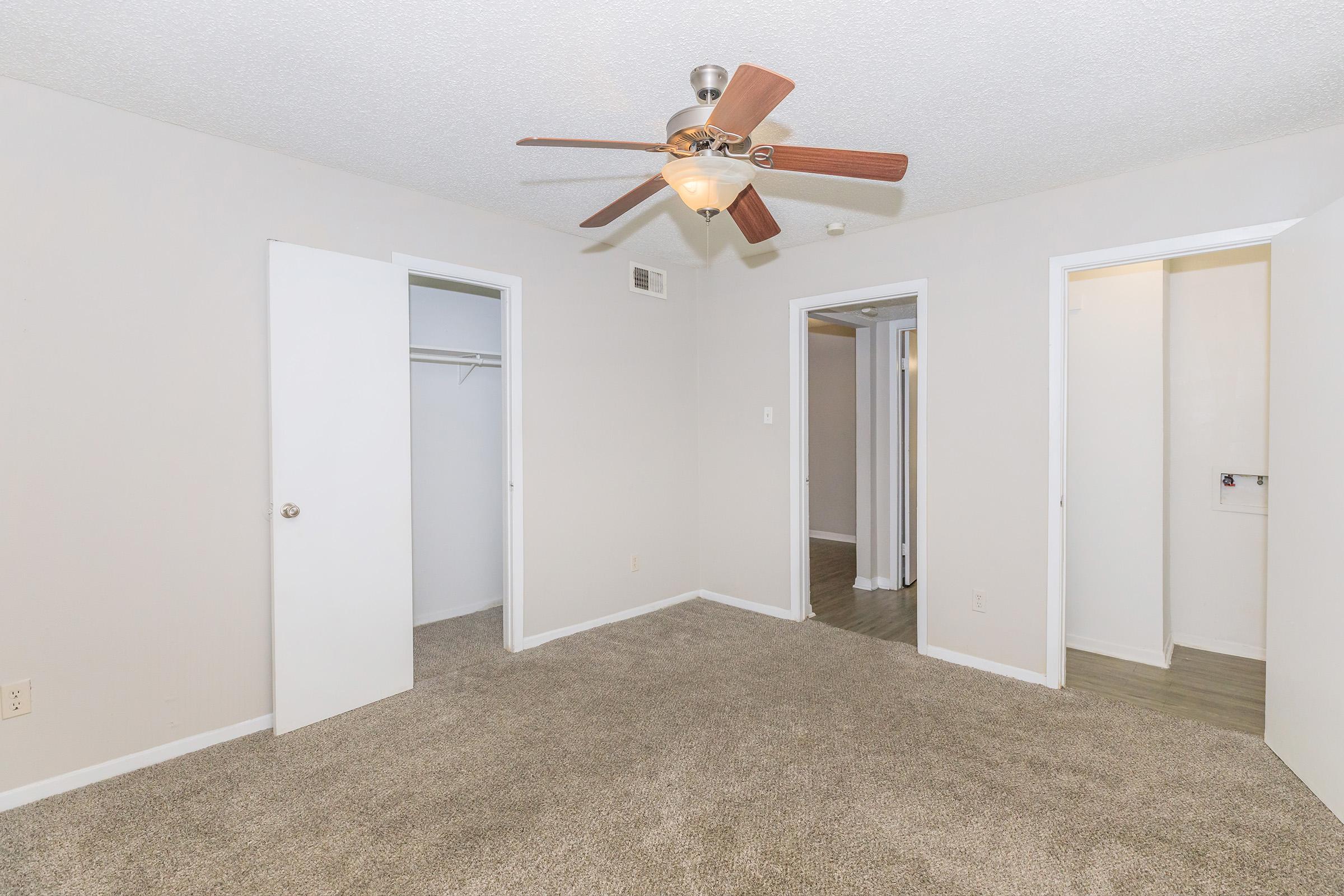 A small, empty room with light beige walls and a ceiling fan. There are two white doors leading to closets on either side. The floor is covered in light-colored carpet, and there is a glimpse of a hallway in the background. The room has a bright, airy feel due to the natural light.