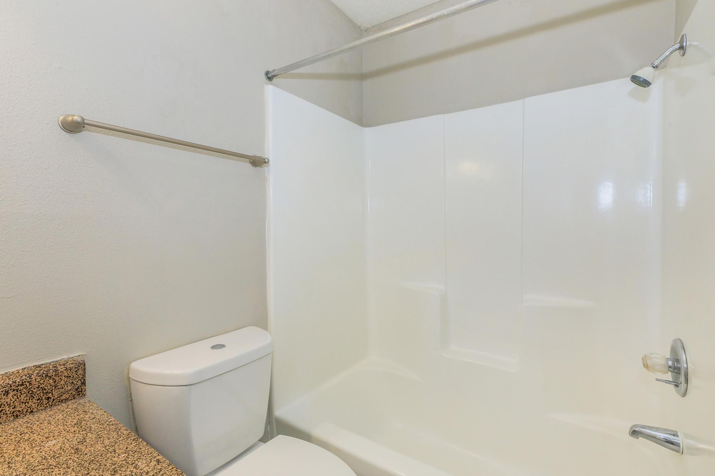 A clean and simple bathroom featuring a white bathtub and shower combo, a modern toilet, and a beige countertop. The walls are painted a light gray, and a stainless steel towel bar is mounted on the wall. The overall look is minimalist and functional, suitable for everyday use.