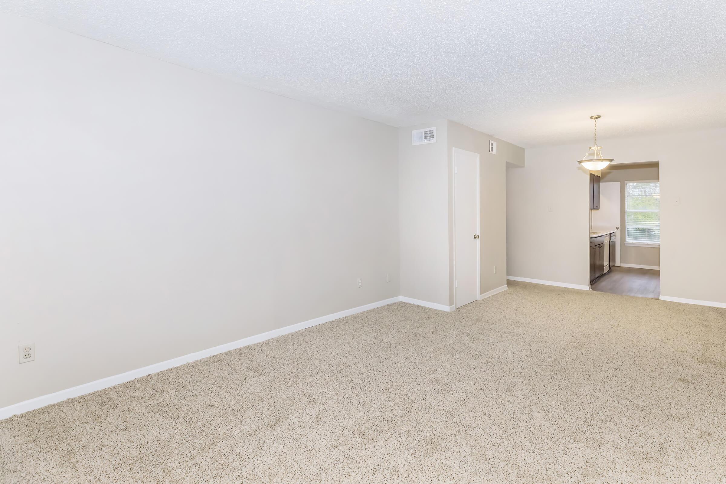 A spacious, empty room with light beige carpet and textured ceiling. The walls are painted in a light color, and there’s a doorway leading to a kitchen area in the background. Natural light comes from a window, and a simple chandelier hangs from the ceiling near the entry to the kitchen.