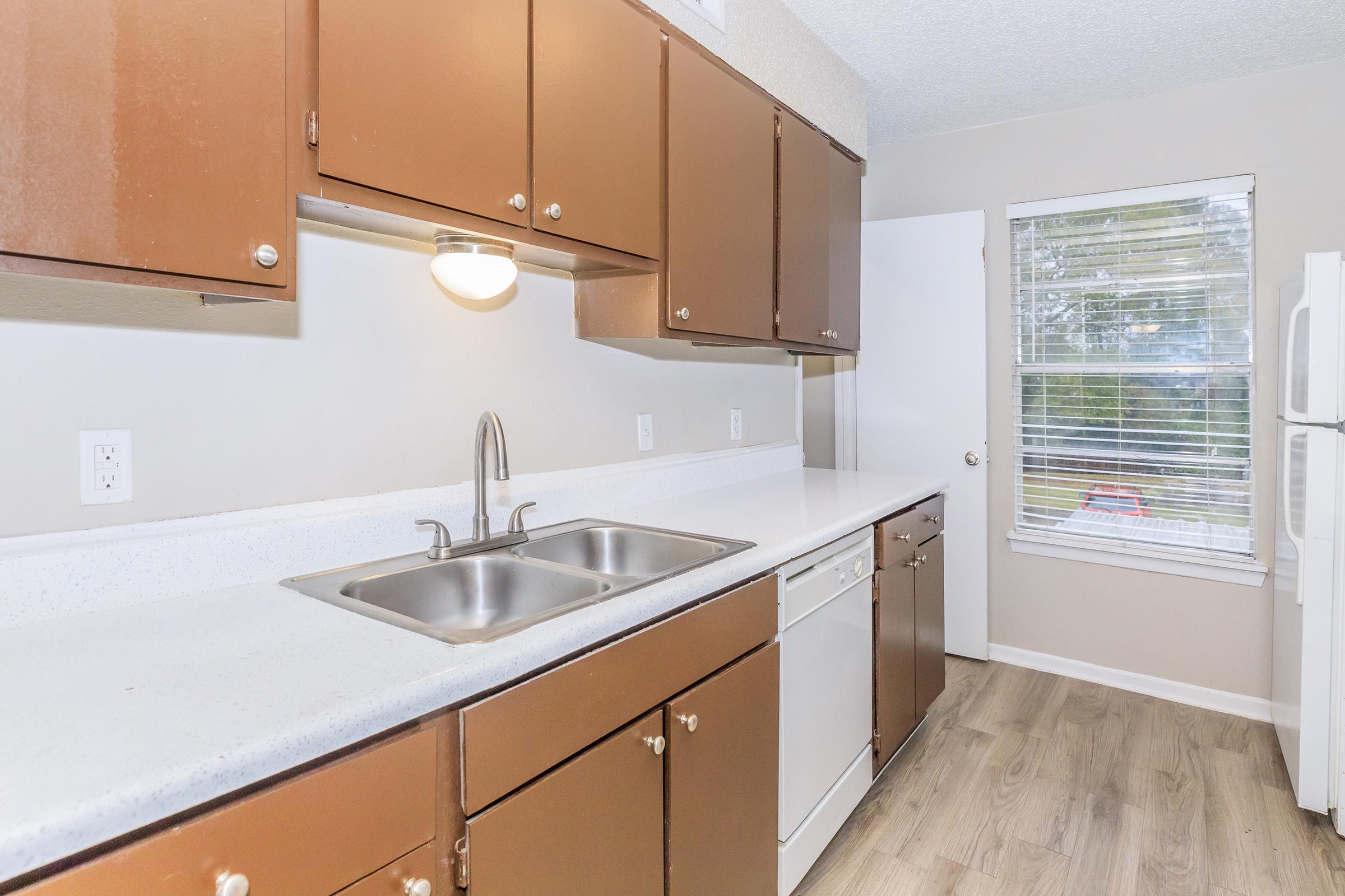 A modern kitchen with brown cabinets, a double sink, and a white dishwasher. The countertops are light-colored. A door leads to an outside area, and there's a window with blinds allowing natural light to enter. The floor is finished with light wood.