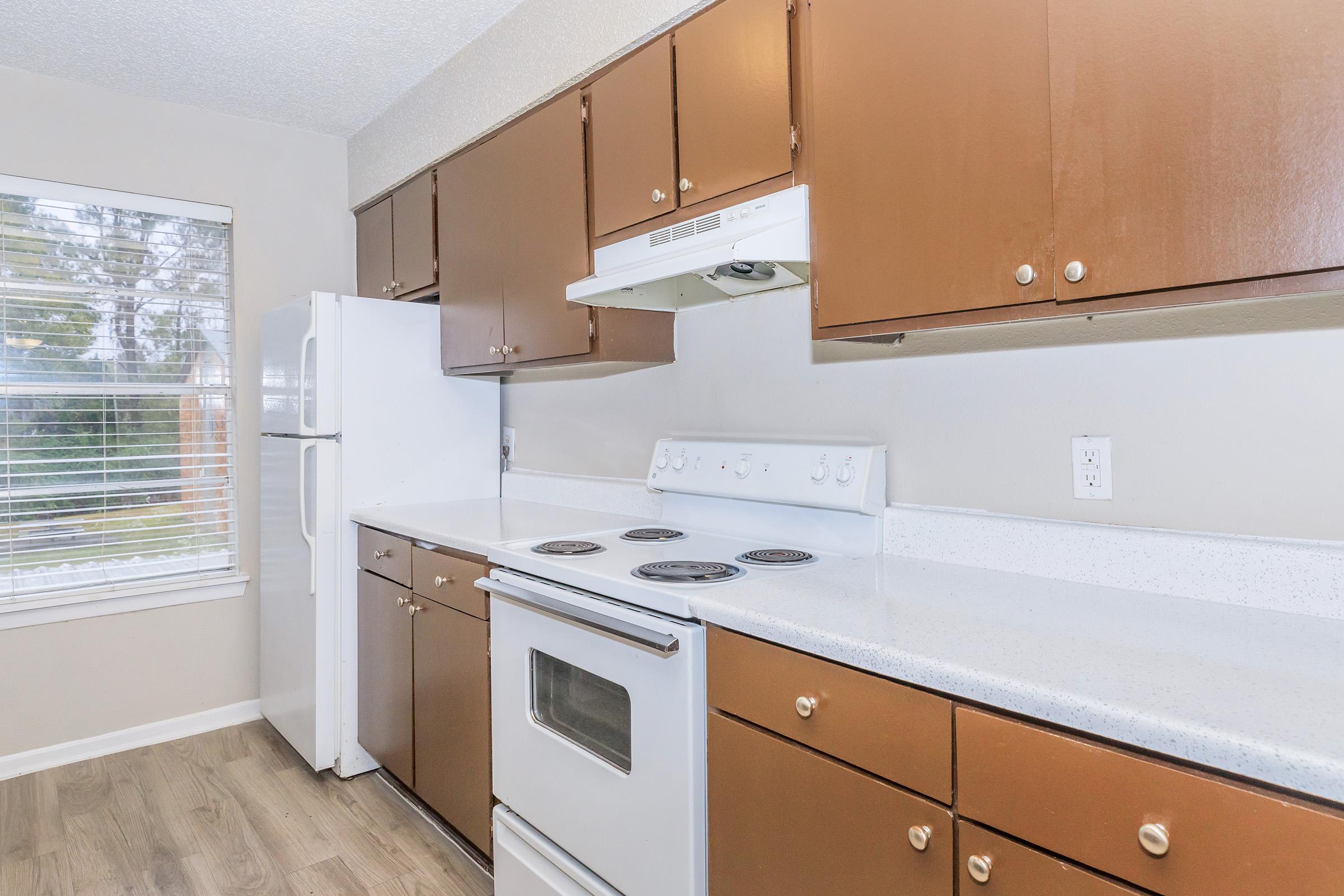 A modern kitchen featuring brown cabinets, a white stove with four burners, and a built-in microwave above. A white refrigerator is positioned next to a window with blinds, allowing natural light to enter. The countertop is light-colored, providing a clean and functional appearance.