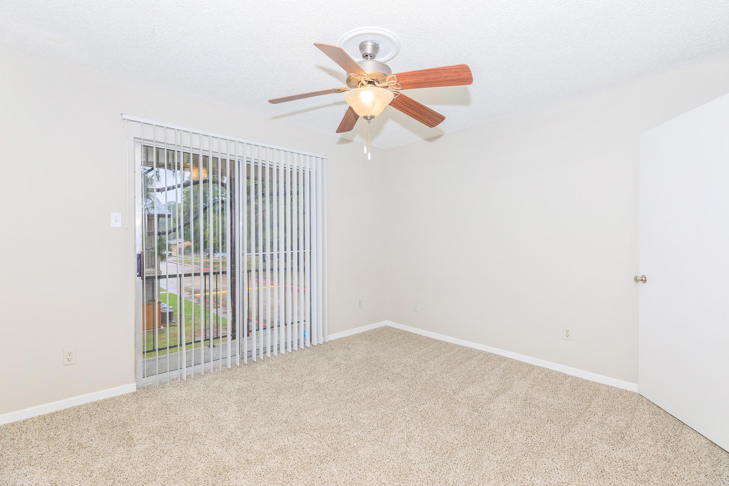 A light, empty room featuring beige carpet and a ceiling fan with wooden blades. A sliding glass door with vertical blinds leads to an outdoor area, with a plain white door visible on the right. The walls are a neutral color, creating a bright and airy atmosphere.