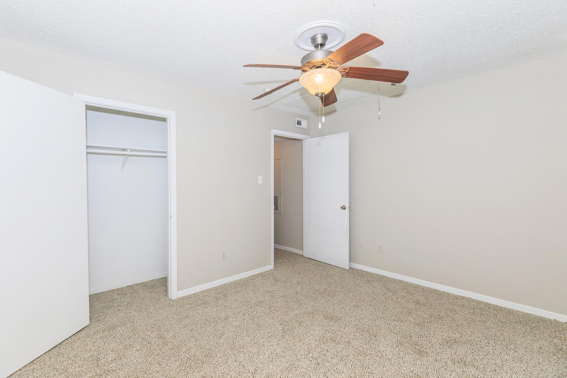 A vacant bedroom with beige carpet and neutral-colored walls. It features a ceiling fan with wooden blades and light fixture, an open closet on the left, and a closed door on the right. The room is well-lit and has minimal furnishings, creating a spacious feel.