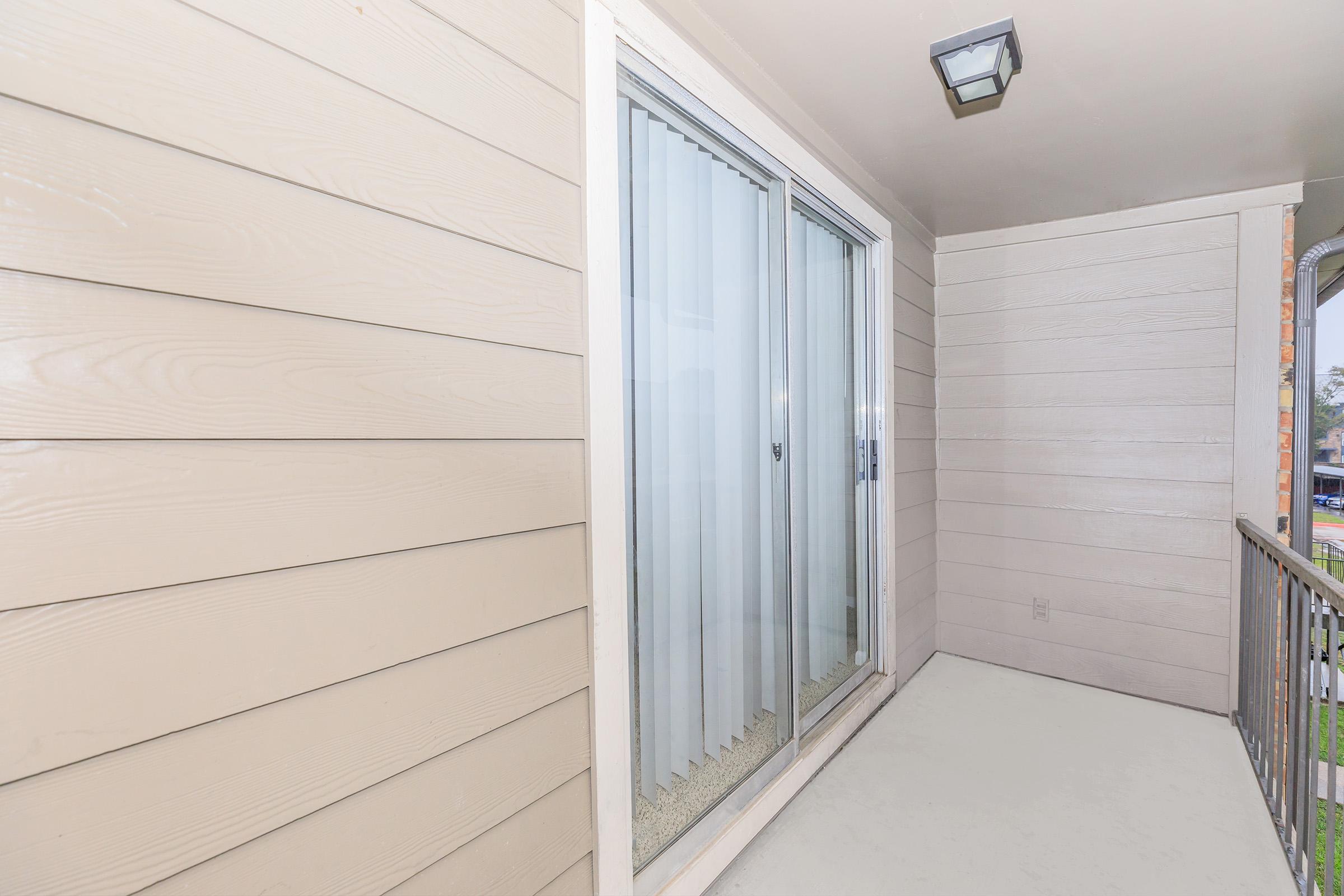 A small balcony featuring sliding glass doors, with beige wooden siding on the walls. The balcony floor is light-colored, and there is a simple light fixture on the ceiling. The view outside is partially visible through the glass doors.