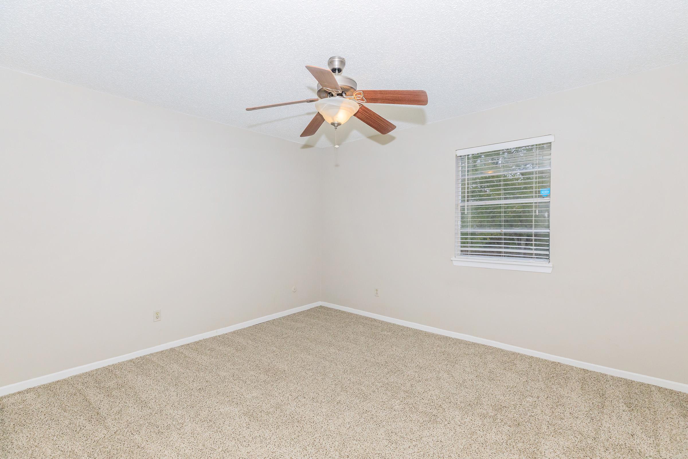 A spacious, empty room with light beige carpet and white walls. A ceiling fan with wooden blades hangs in the center. A window with white blinds allows natural light to enter the space. The overall ambiance is bright and uncluttered, ideal for personalization.