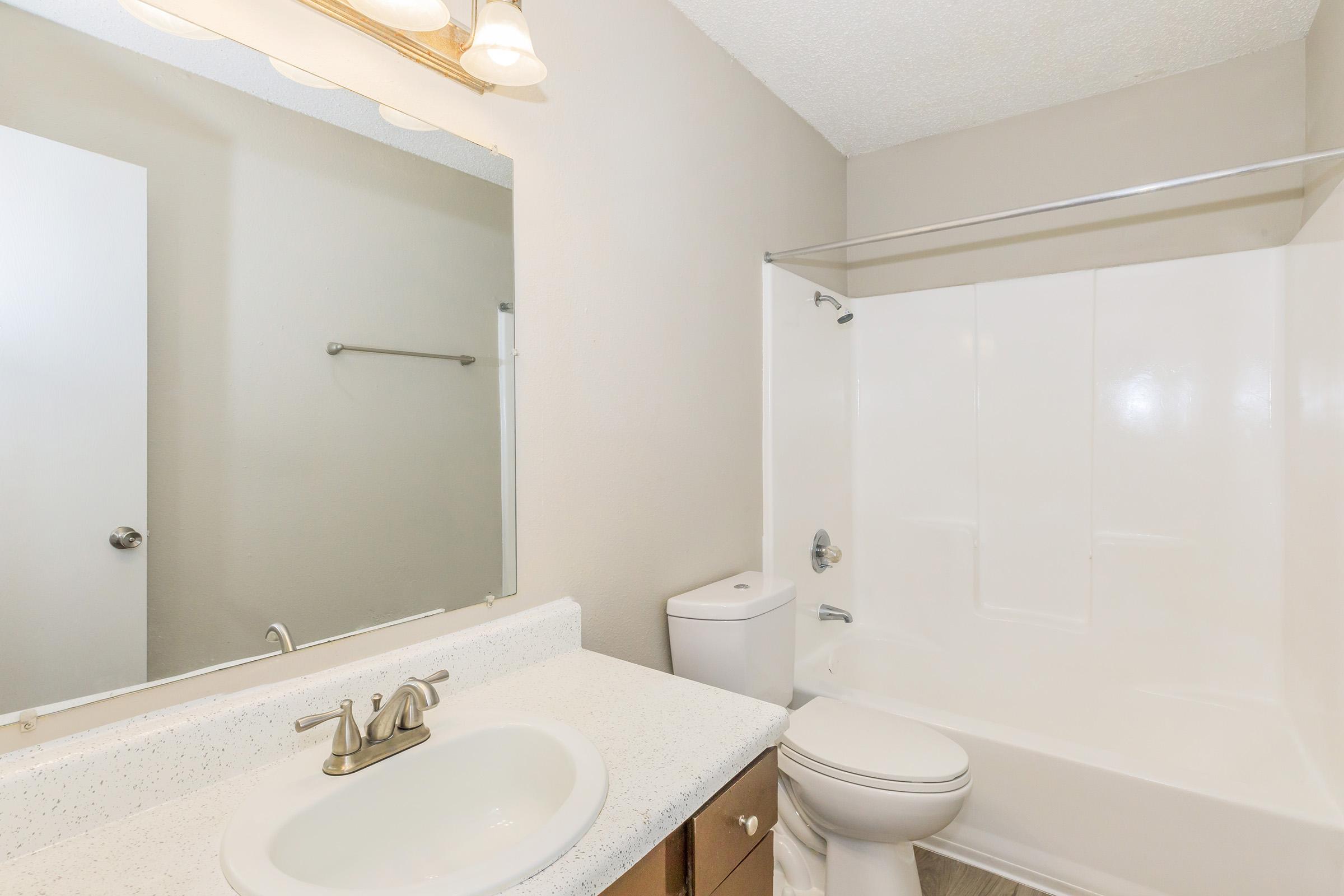 A clean, modern bathroom featuring a white bathtub and shower combination, a light-colored countertop sink with a faucet, a mirror above the sink, and a simple light fixture. The walls are painted a soft neutral color, and a towel rack is mounted on the wall.